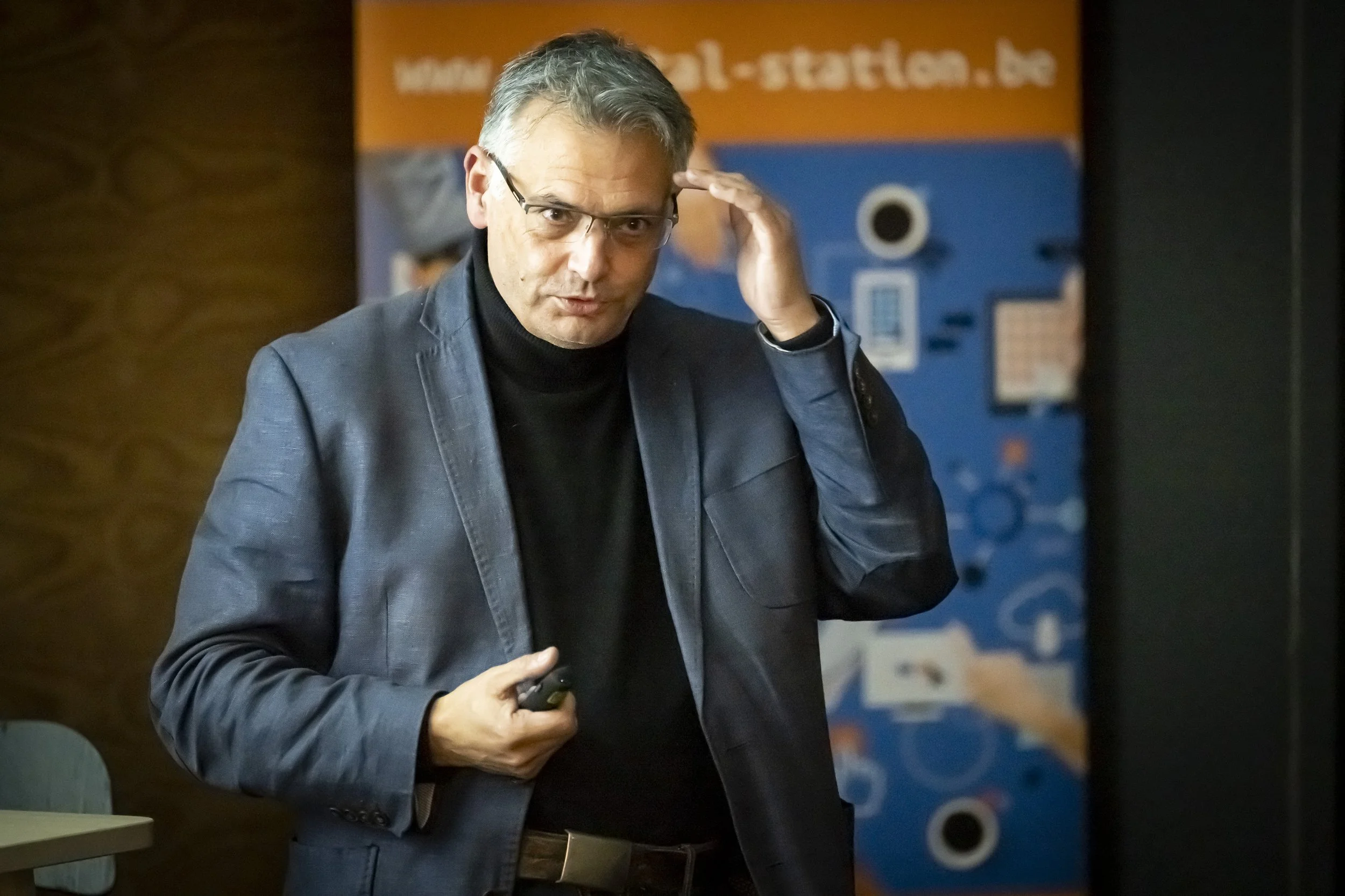 Vincent Parissis, en veste bleue et col roulé noir, porte une main à ses lunettes lors du meetup WalChain à Charleroi.