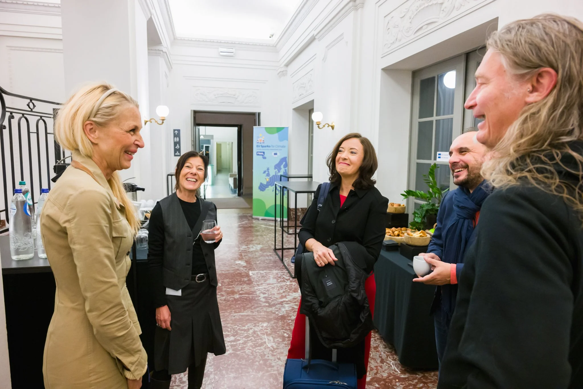 Photo pendant un événement à Bruxelles. Plusieurs personnes souriantes discutent debout dans un espace intérieur élégant, avec des boissons et de la nourriture sur une table en arrière-plan.