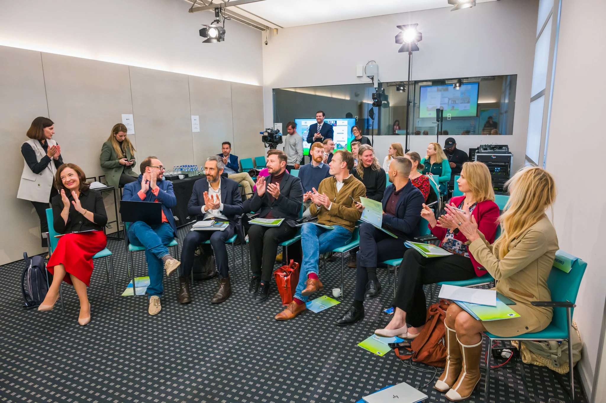 Groupe de personnes applaudissant lors d'un événement en salle, avec des chaises et du matériel audiovisuel. La photo a été réalisée dans le contexte d'une conférence à Bruelles.