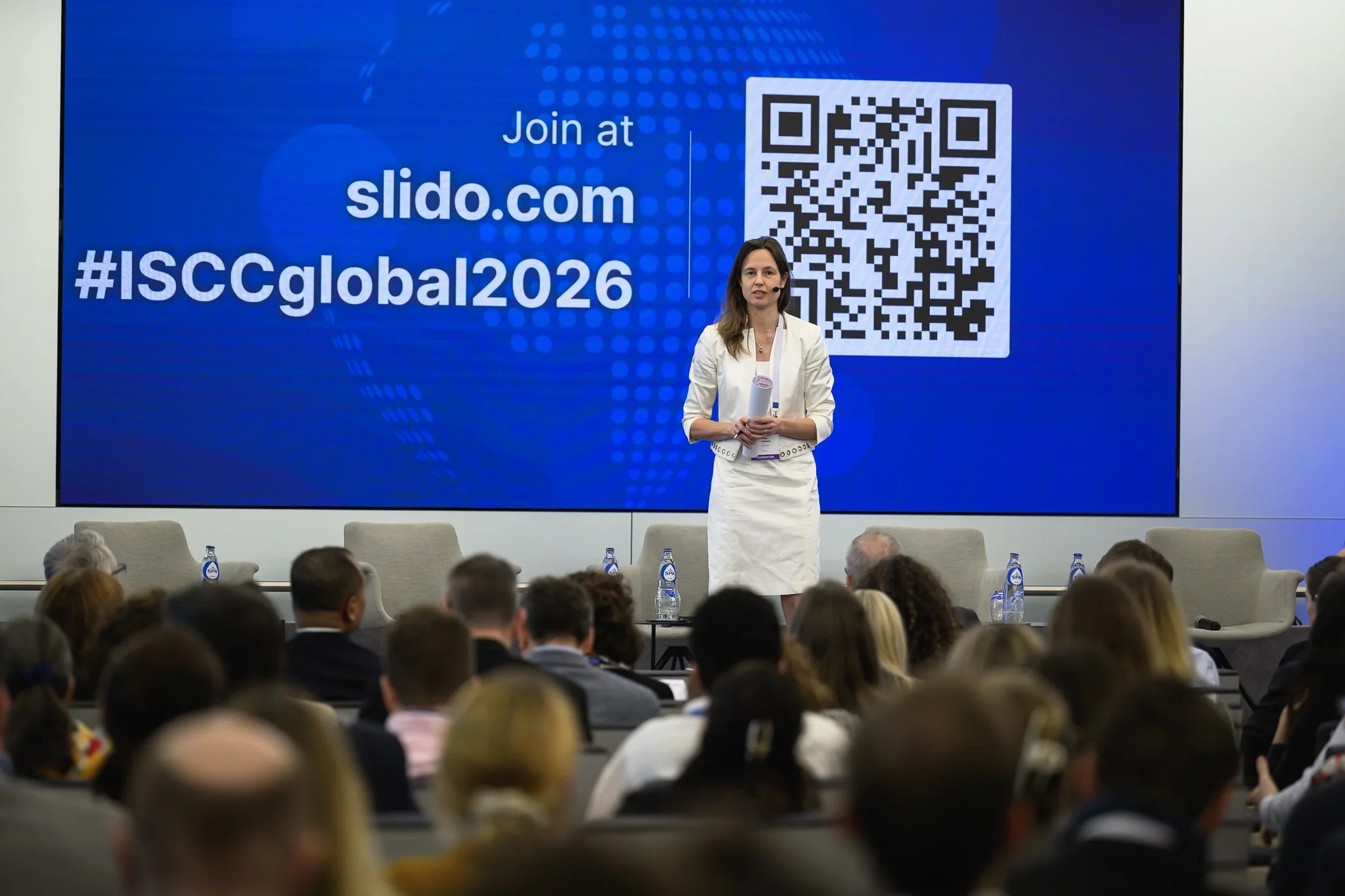 A woman stands on stage beside a screen displaying a Slido address and QR code.