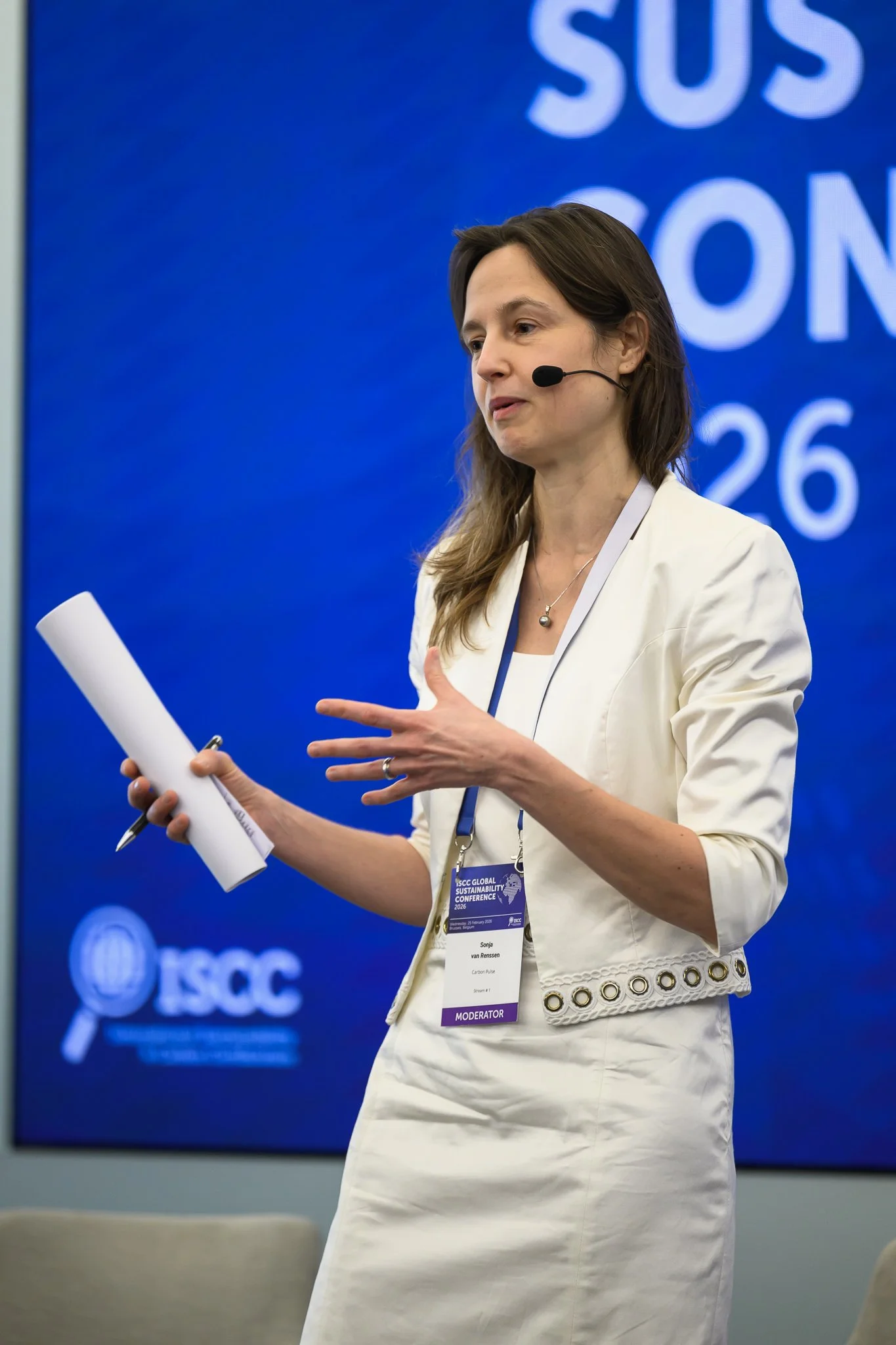 A woman wearing a white outfit and headset microphone addresses the conference while holding notes.