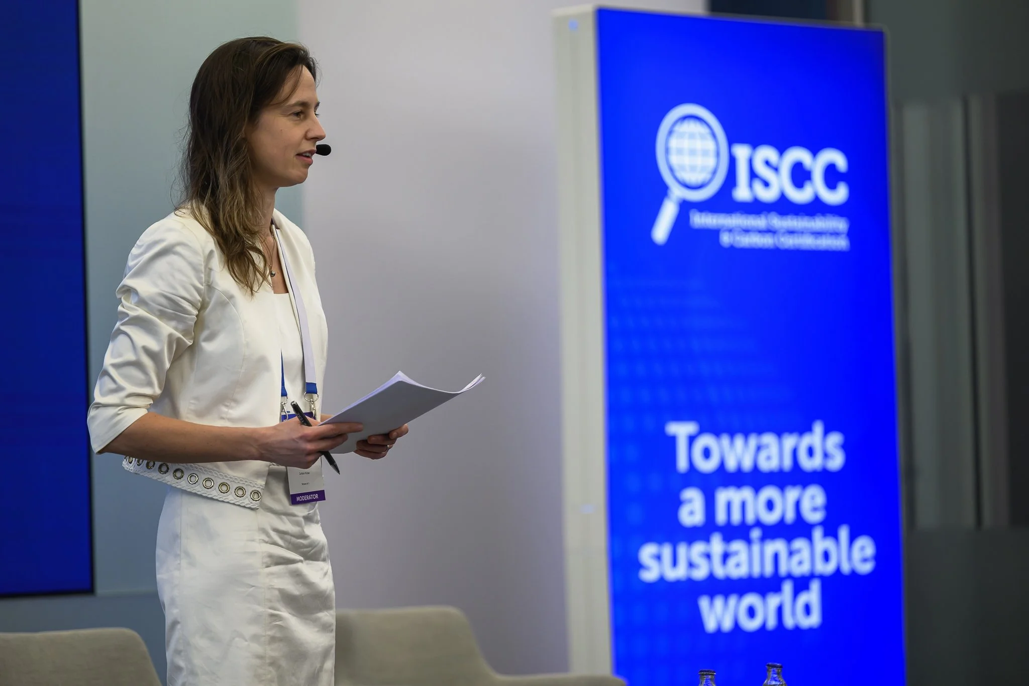 A woman in a white outfit holds notes beside an ISCC banner reading Towards a more sustainable world.