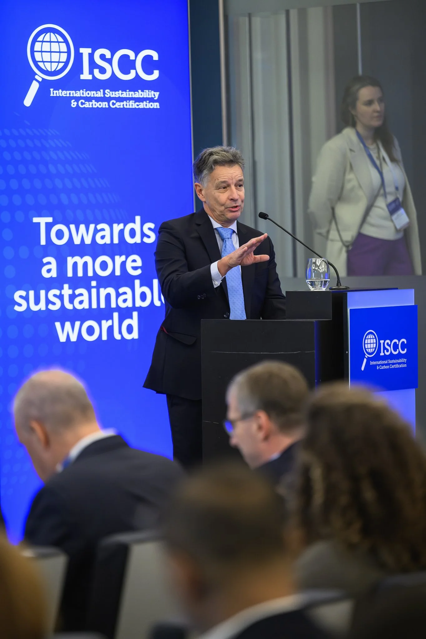 A man in a dark suit speaks at a lectern beside a blue ISCC banner.