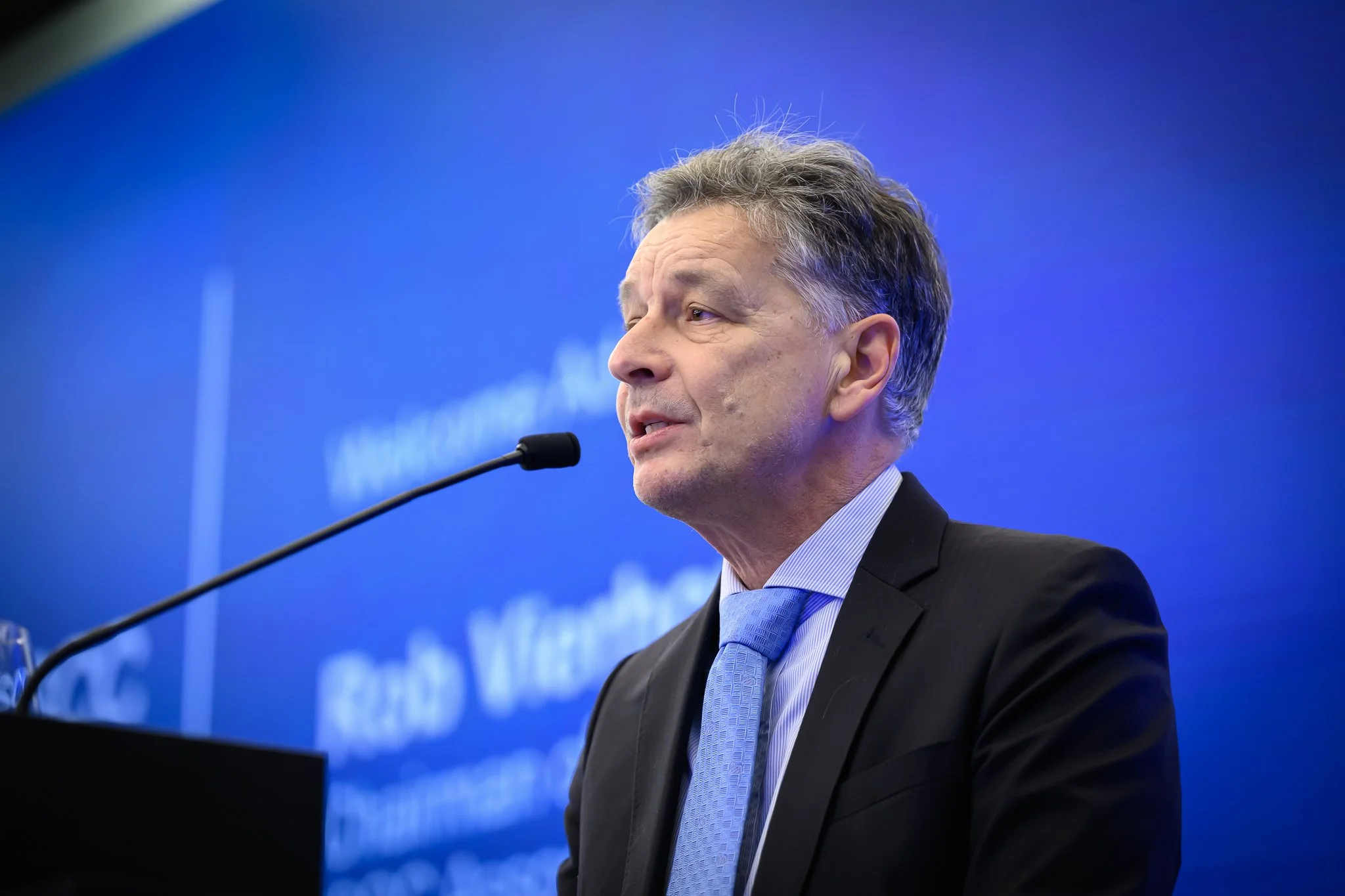 A man in a dark suit and blue tie speaks into a lectern microphone against a blue background.