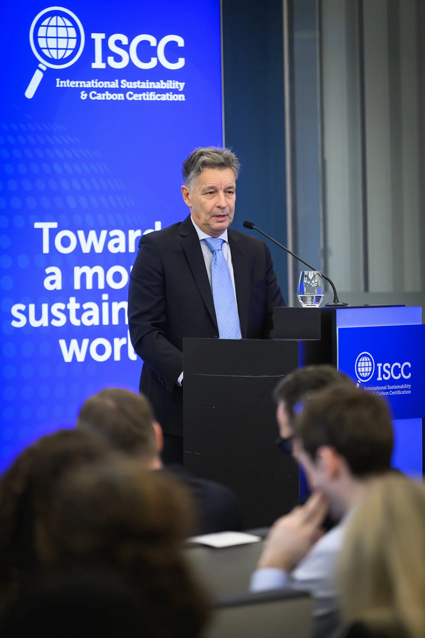 A man speaks from a lectern beside an ISCC banner while the audience listens.
