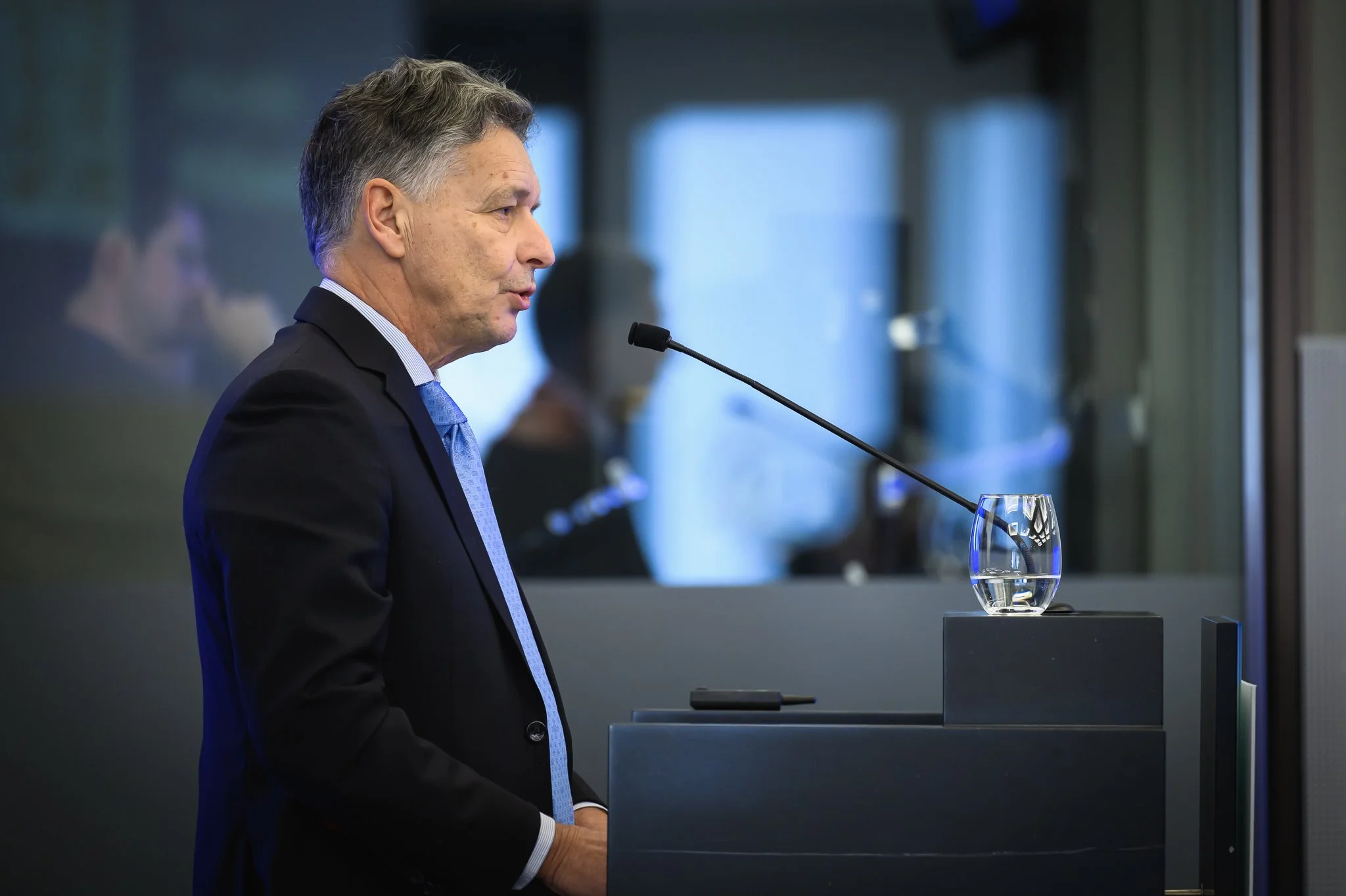 Side view of a man in a dark suit speaking into a lectern microphone.