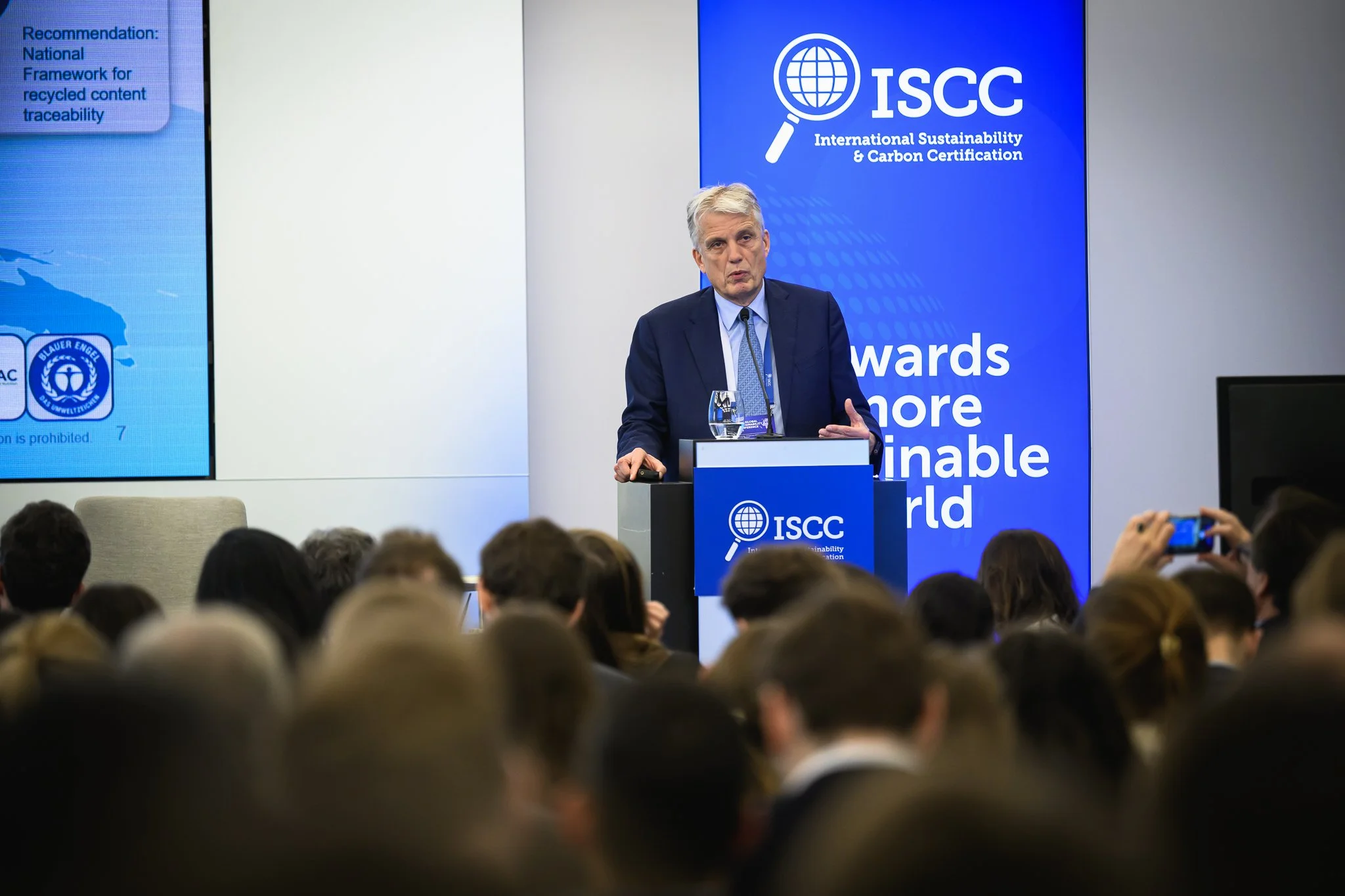 A man in a suit stands at the lectern beside a blue ISCC banner.