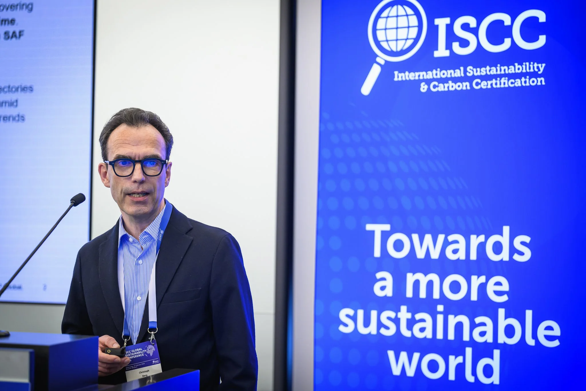 A man with dark-framed glasses stands beside a lectern and an ISCC banner.