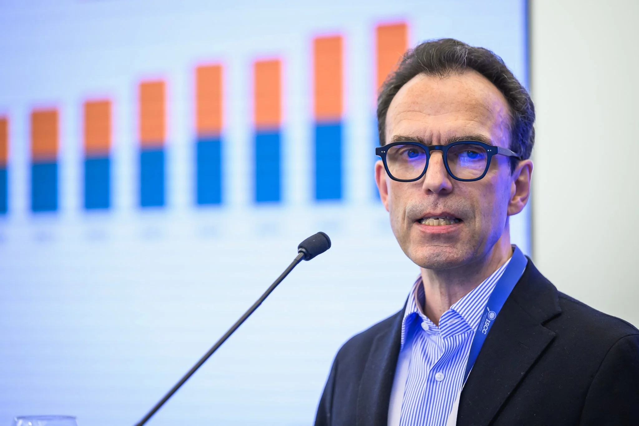 Close-up of a man with glasses speaking at a lectern in front of a bar chart.