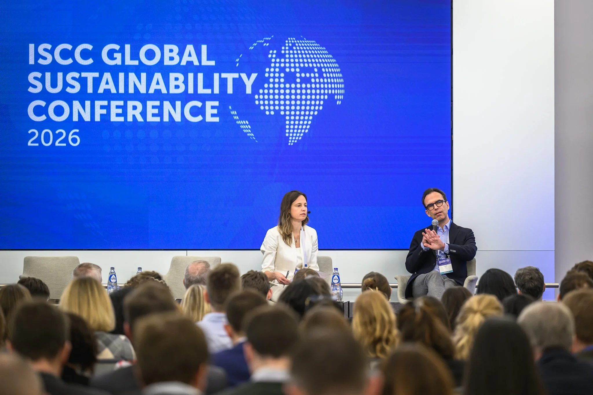 A woman and a man sit on stage for a discussion in front of the blue ISCC backdrop.