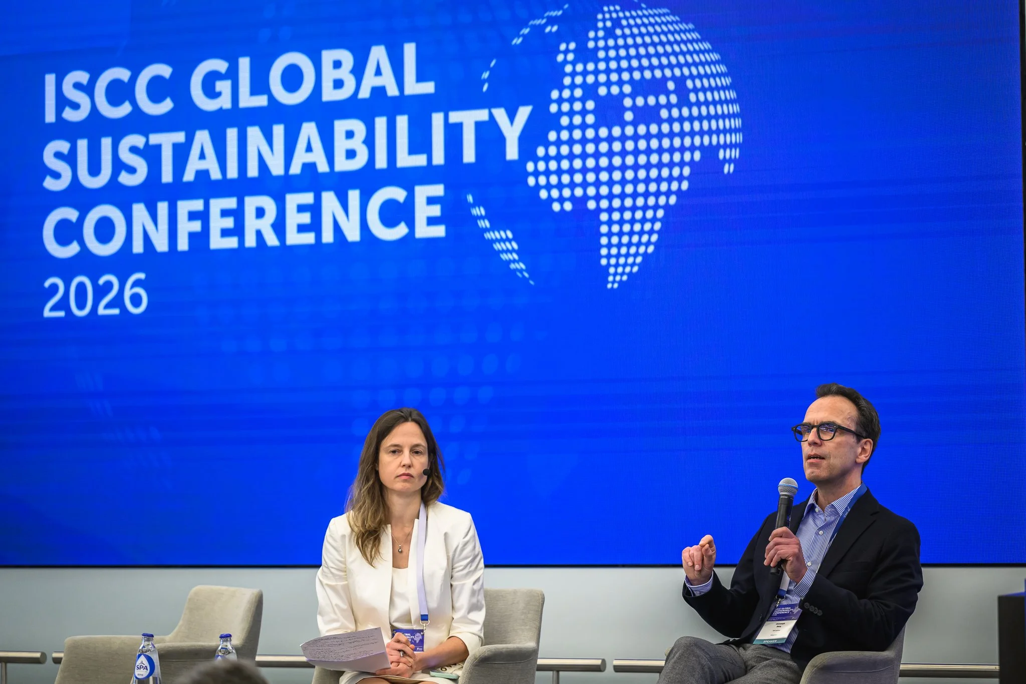 A woman listens as a seated man speaks into a microphone on the conference stage.