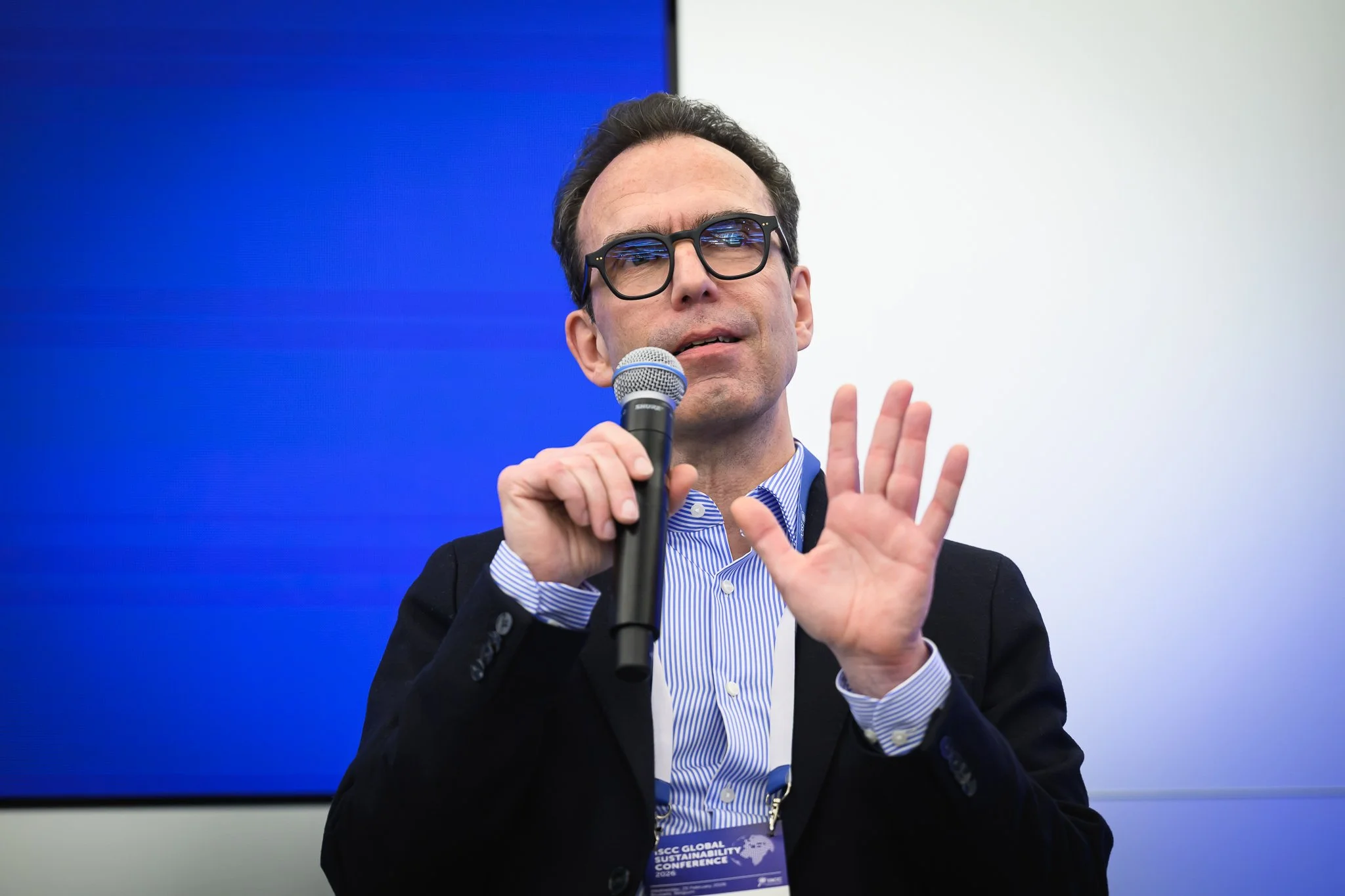 A man with glasses speaks into a handheld microphone and raises an open hand.