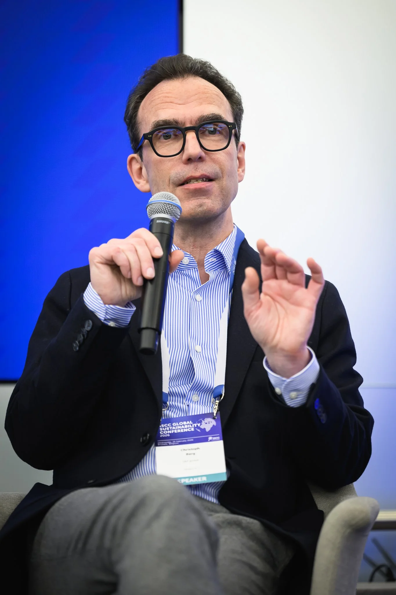 A seated man with glasses gestures while holding a microphone.