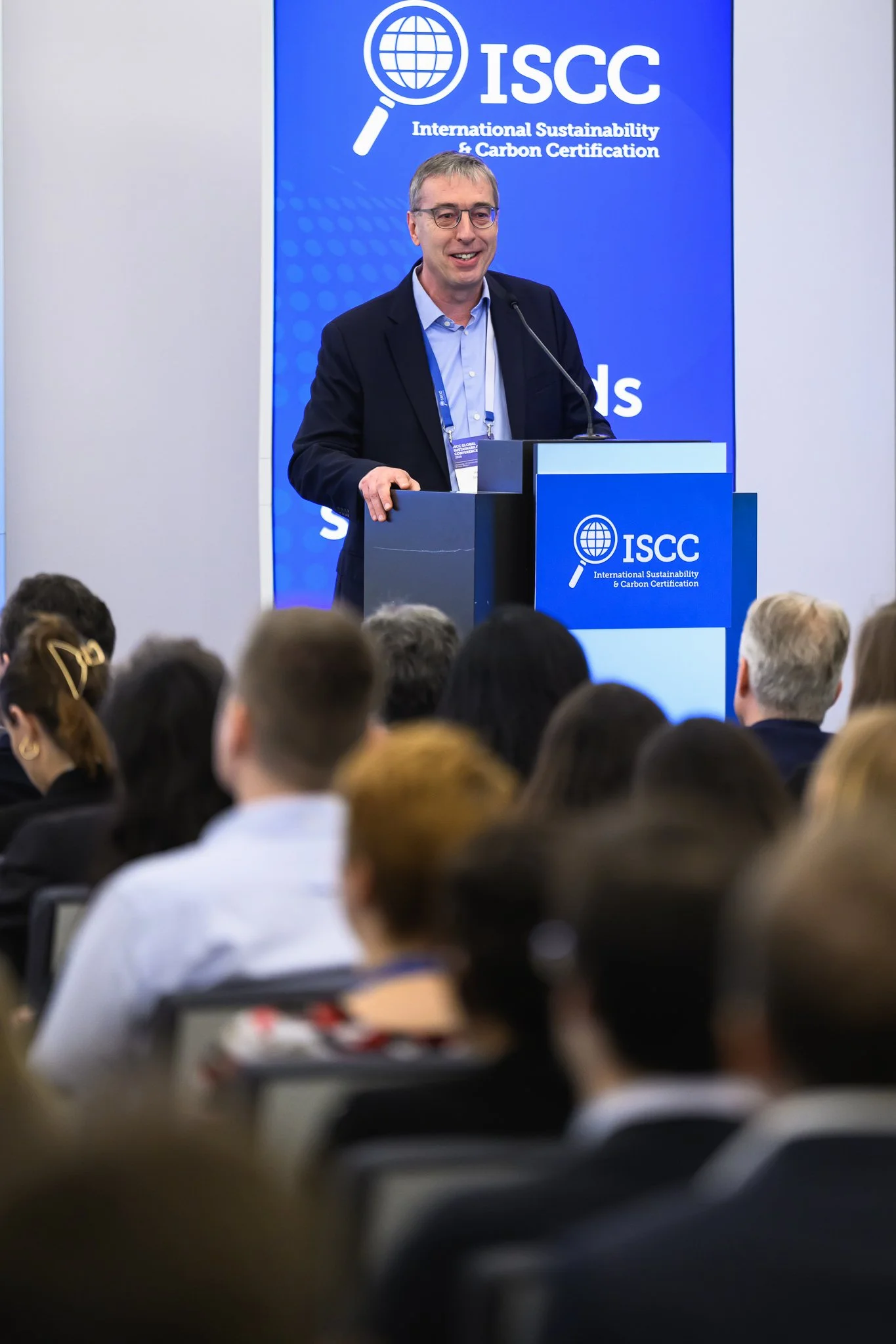 A speaker at the lectern addresses rows of seated conference participants.