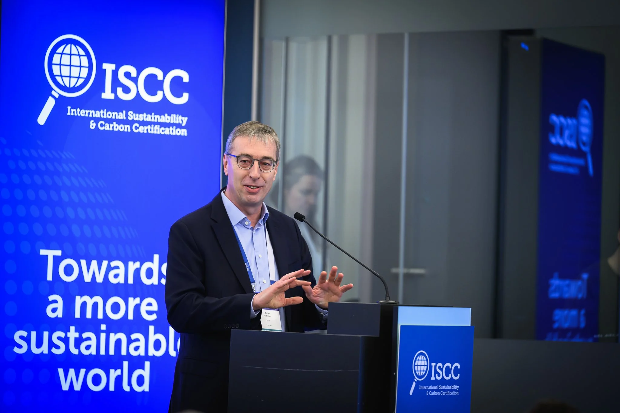 A man with glasses gestures while presenting at a lectern beside an ISCC banner.