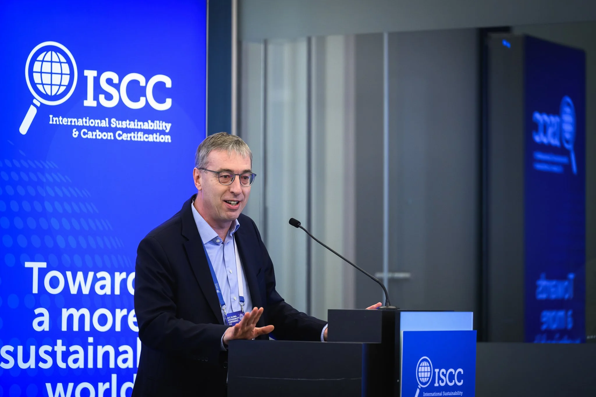 A speaker gestures with one hand beside a blue ISCC banner.