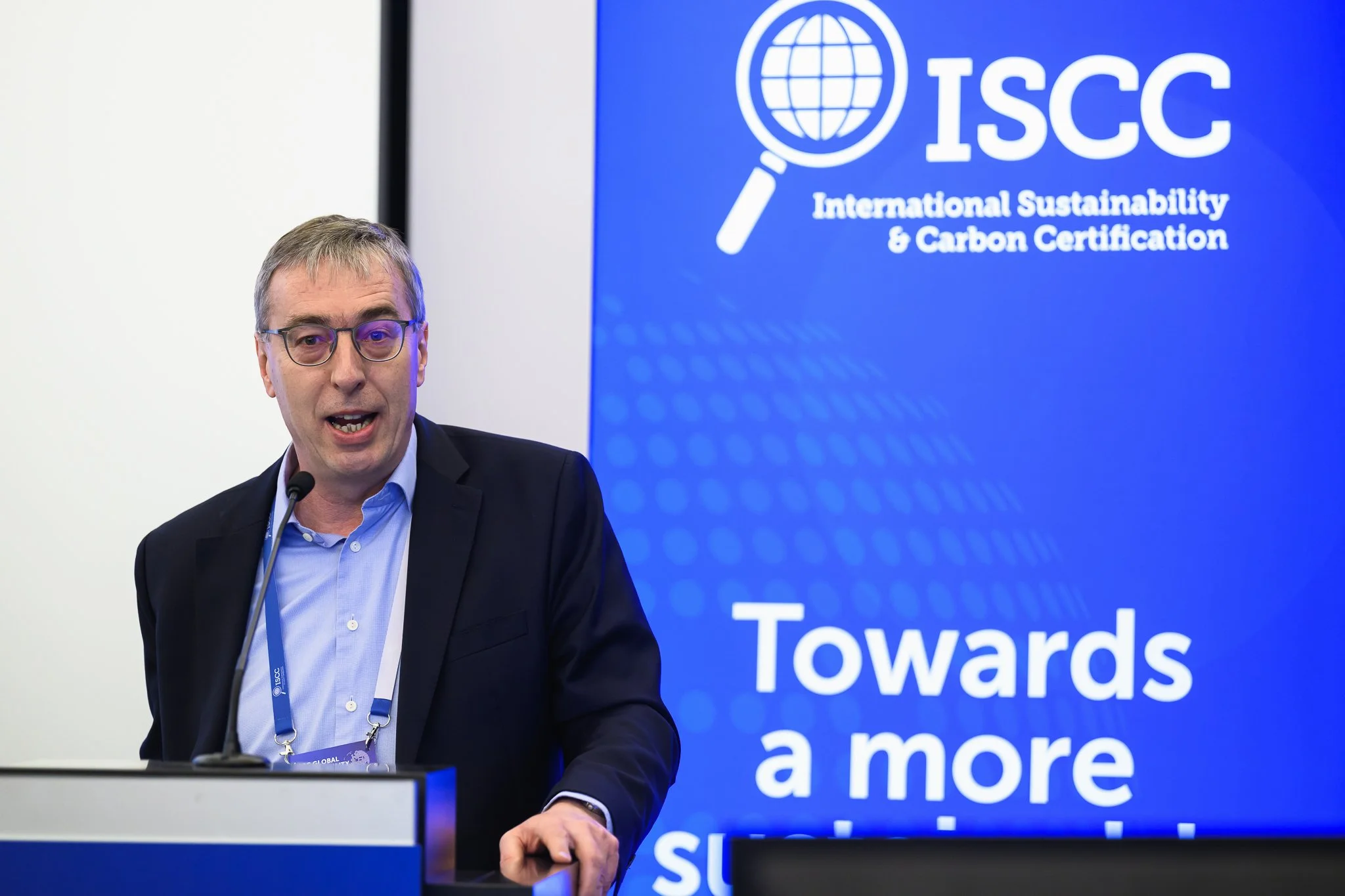 A man with glasses addresses the conference from behind a lectern.
