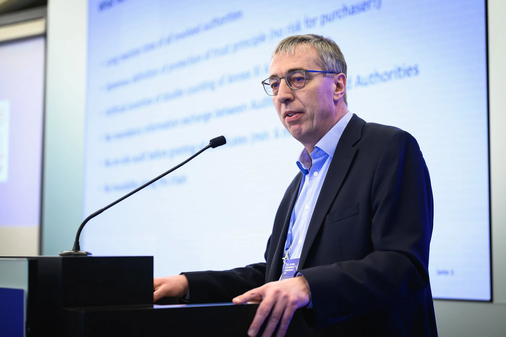A man with glasses speaks at a lectern in front of a projected text slide.