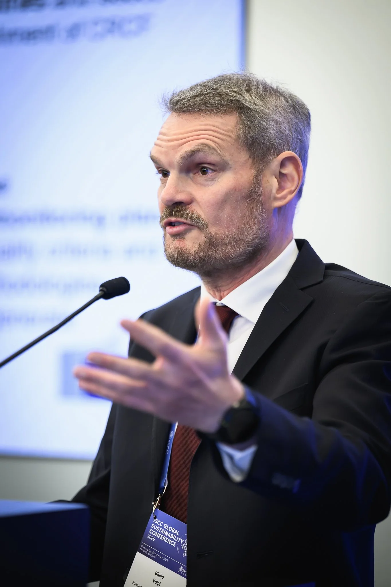 A bearded man in a dark suit gestures while speaking at a lectern.