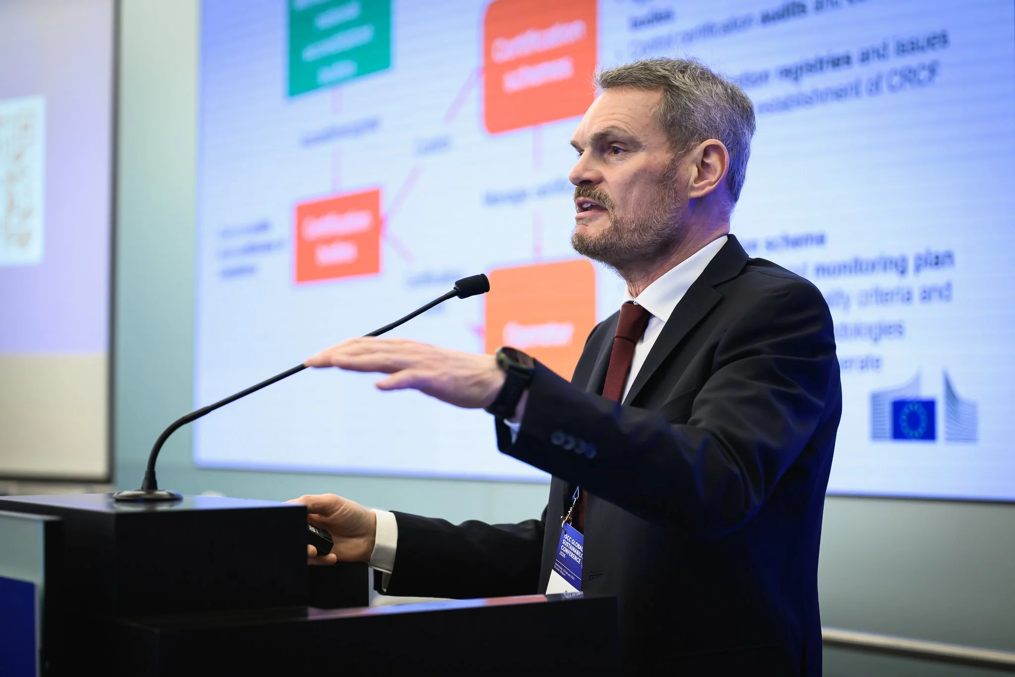 A bearded speaker gestures above the lectern in front of a diagram slide.
