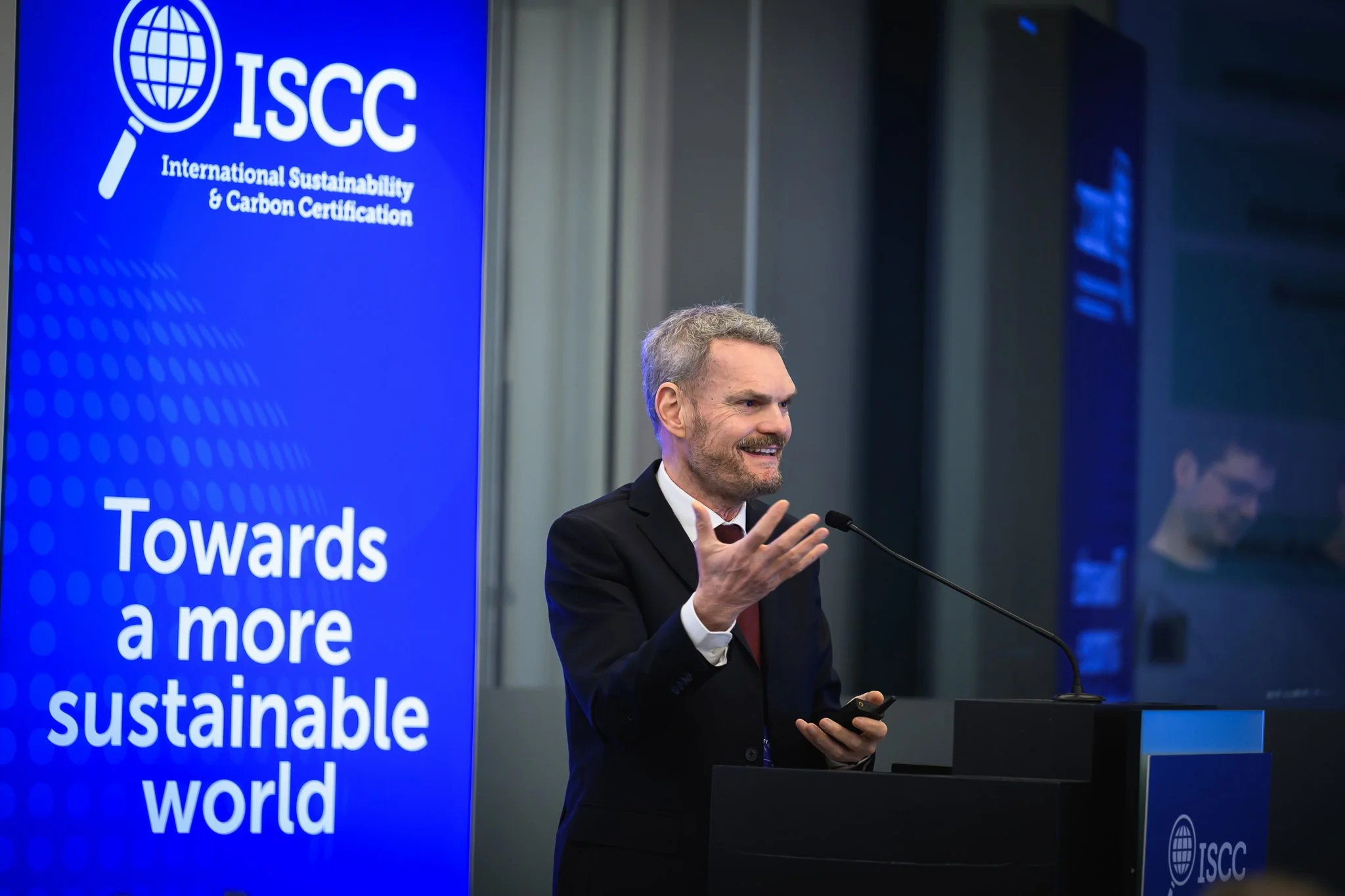 A bearded speaker smiles and raises one hand beside the ISCC banner.