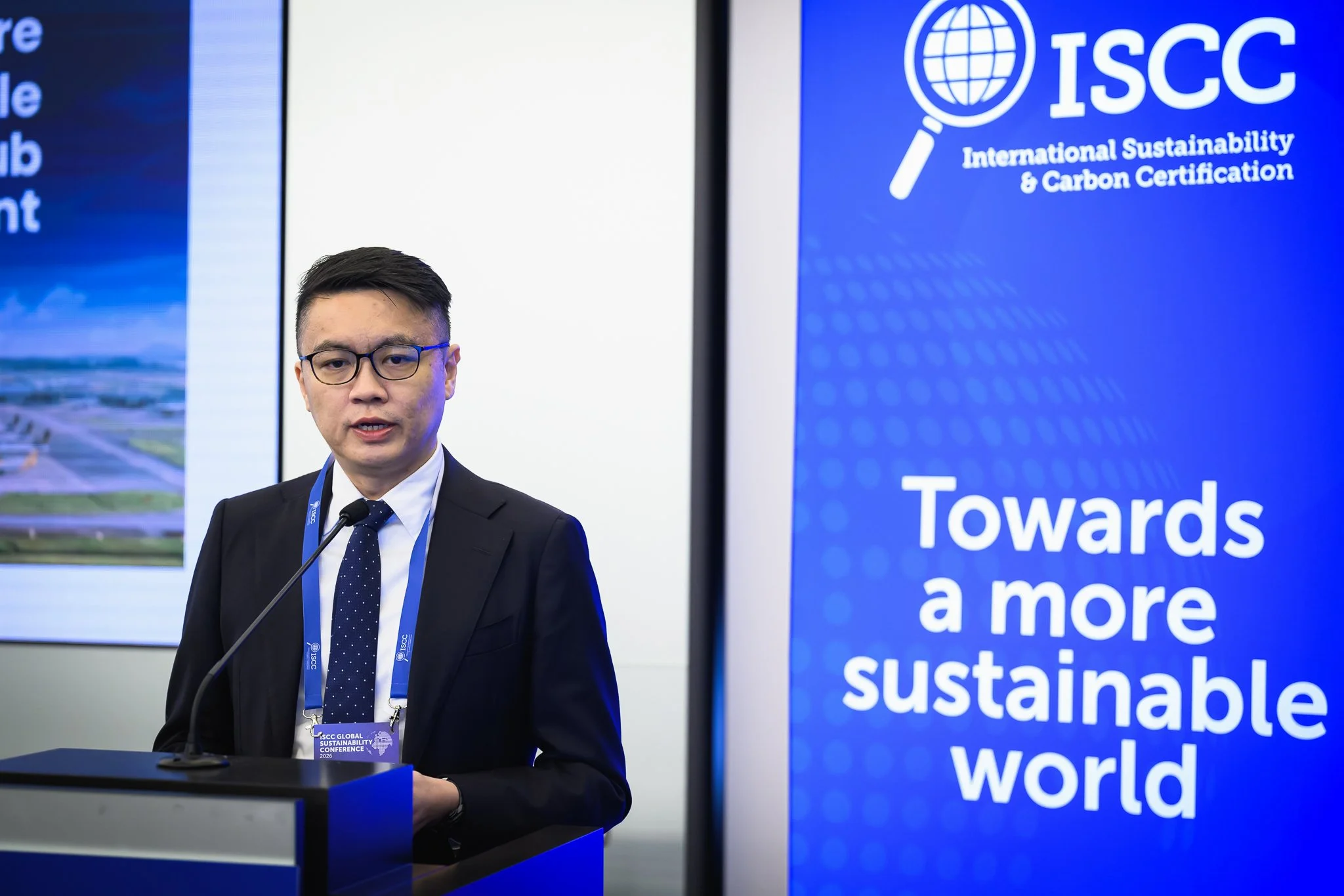 A man in glasses stands at the lectern beside a blue ISCC banner.