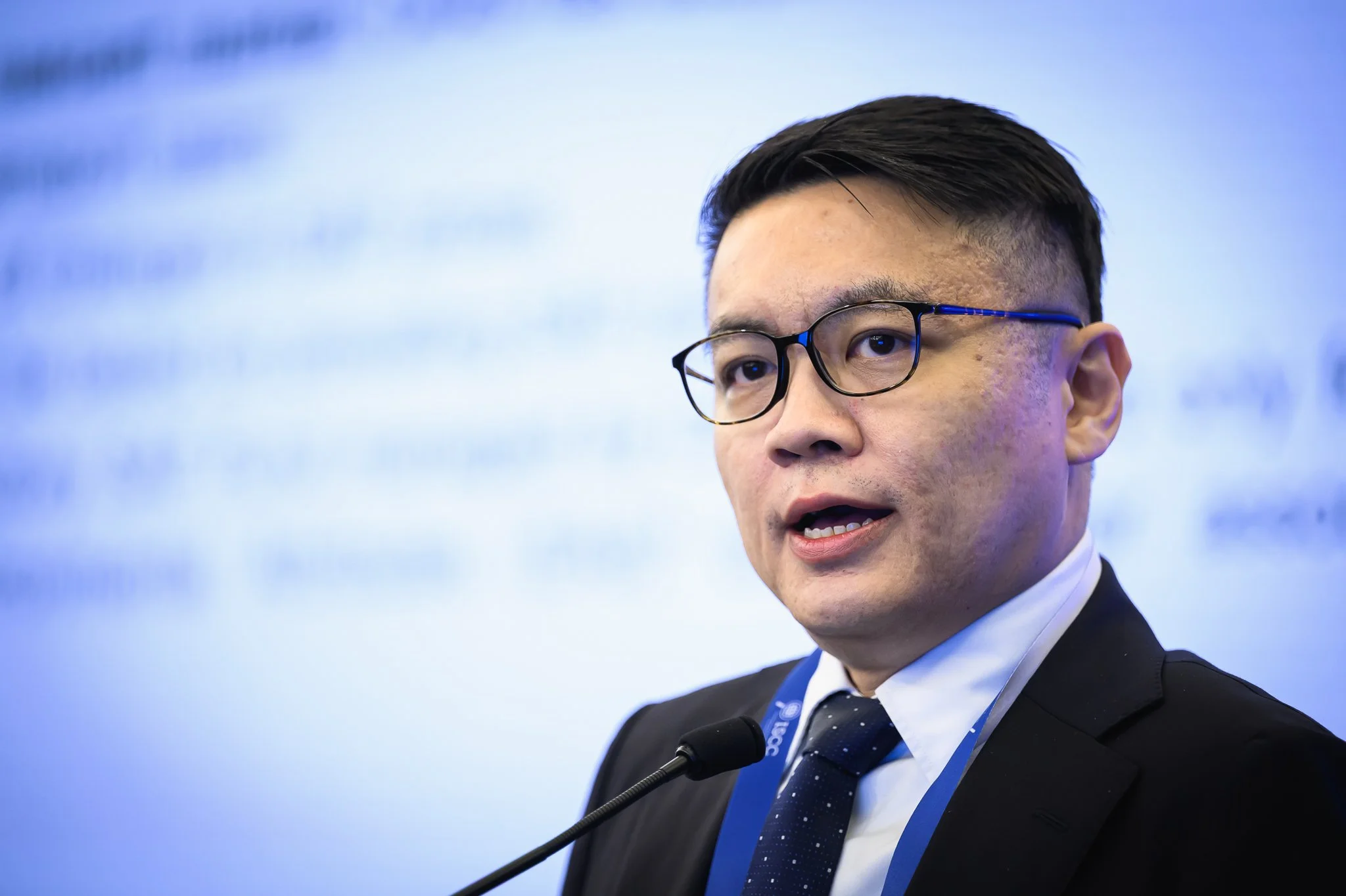 Close-up of a man in glasses speaking into a lectern microphone.