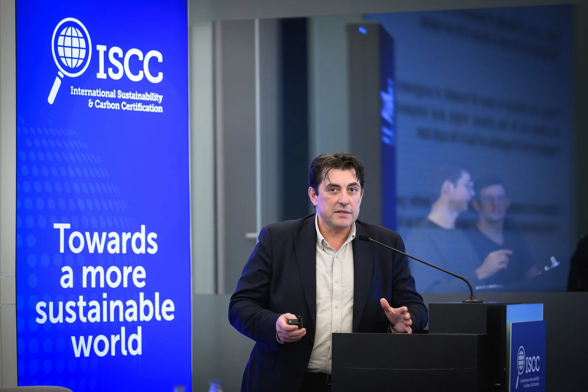A man holds a presentation remote and gestures at a lectern beside an ISCC banner.