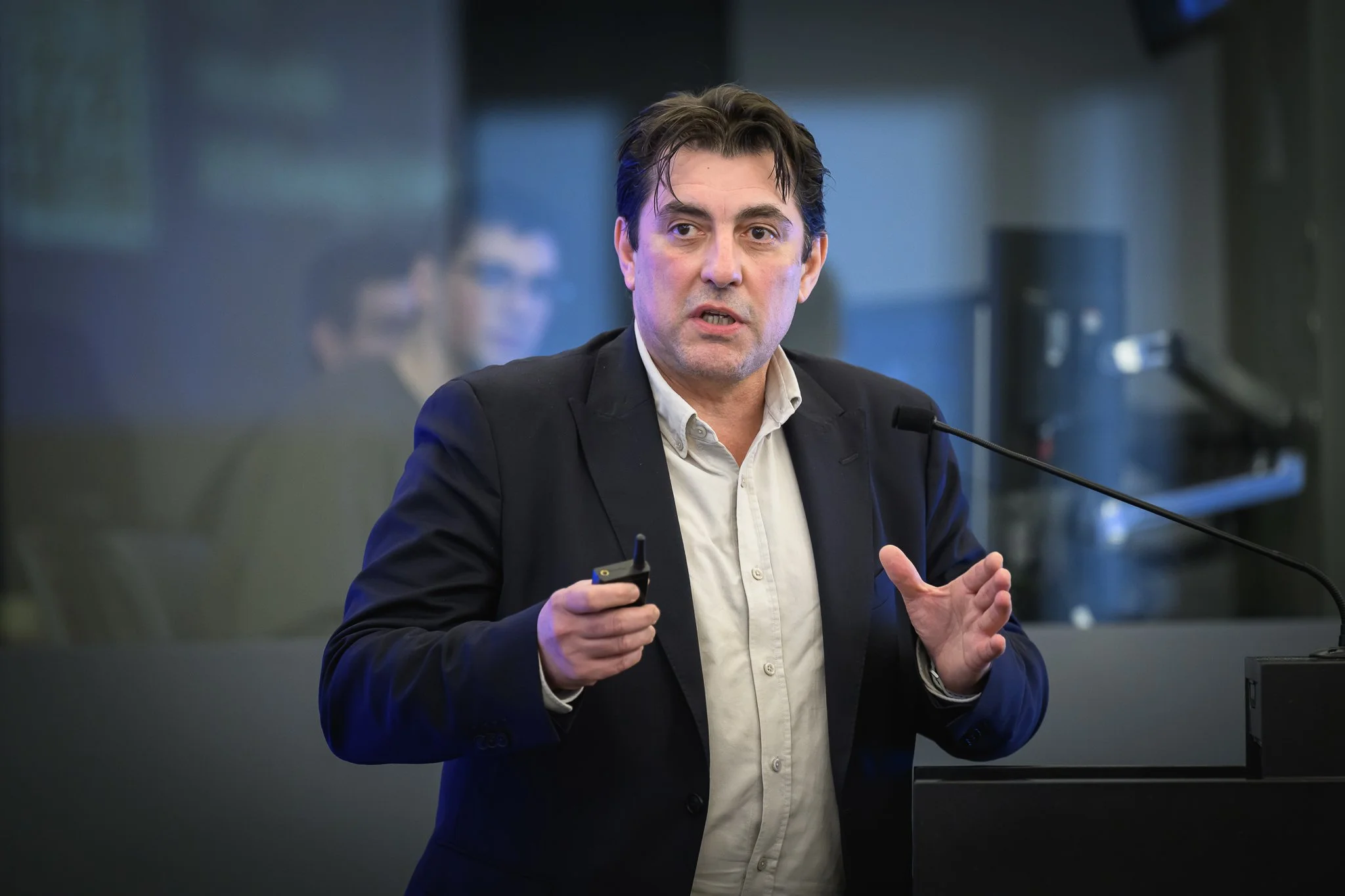 A man gestures with one hand while presenting beside the lectern.