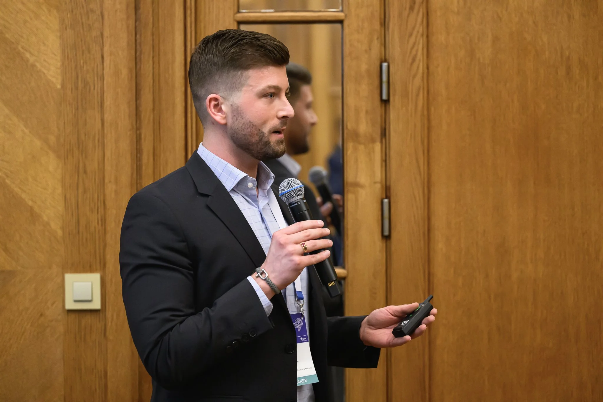 A bearded man holds a microphone and presentation remote beside a wooden wall.