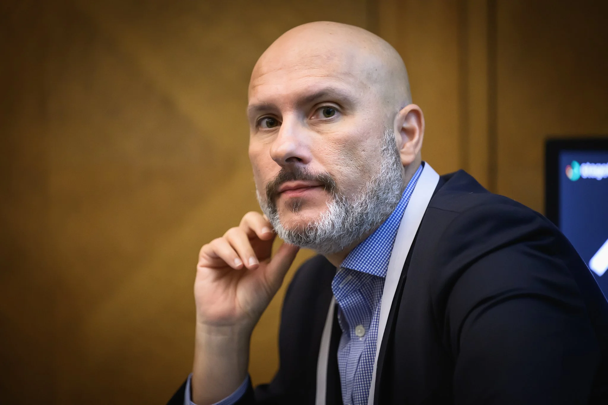 Close-up of a seated bald man with a grey beard listening with one hand near his ear.