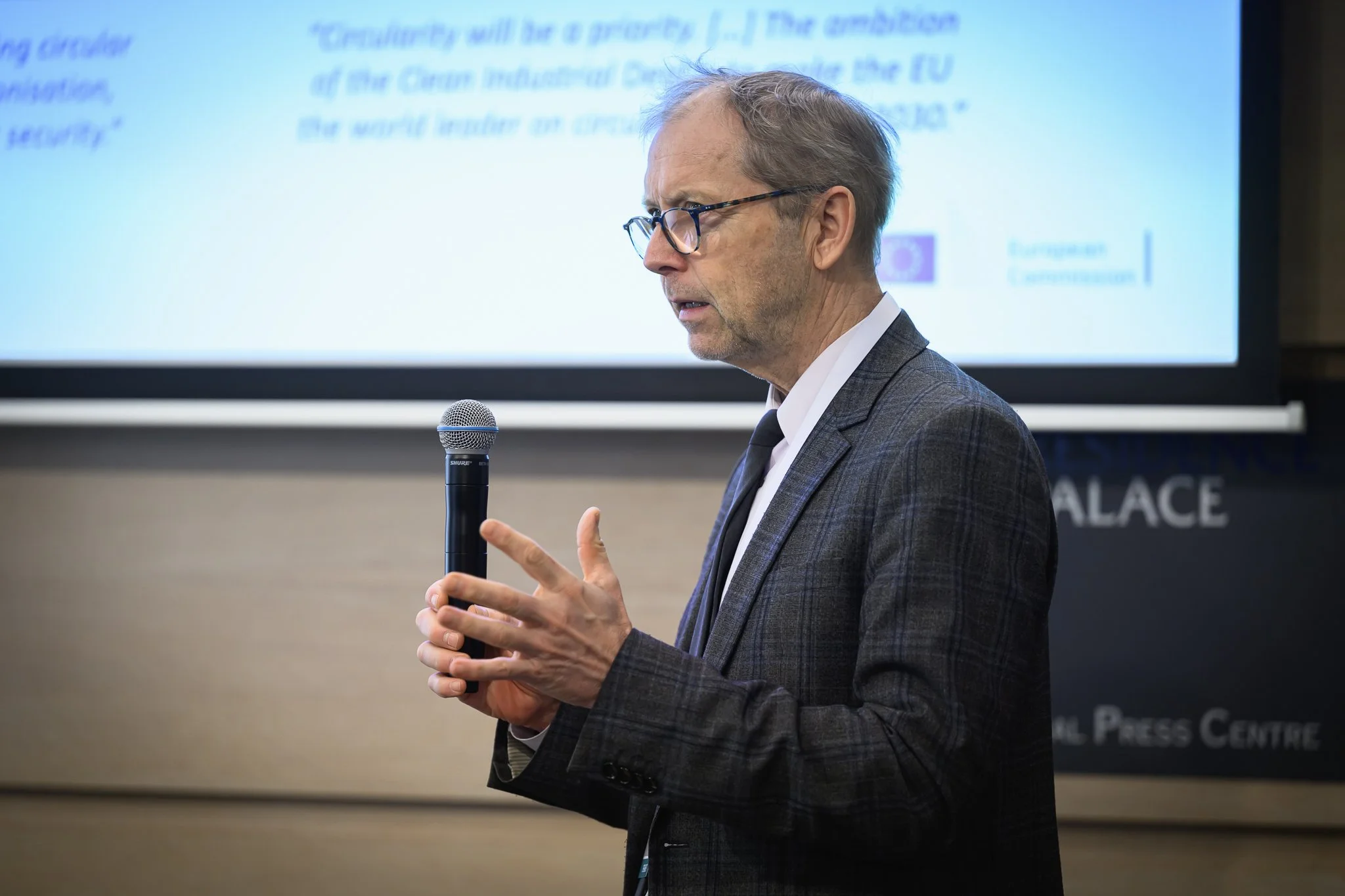 A man with glasses speaks into a handheld microphone in front of a projected slide.