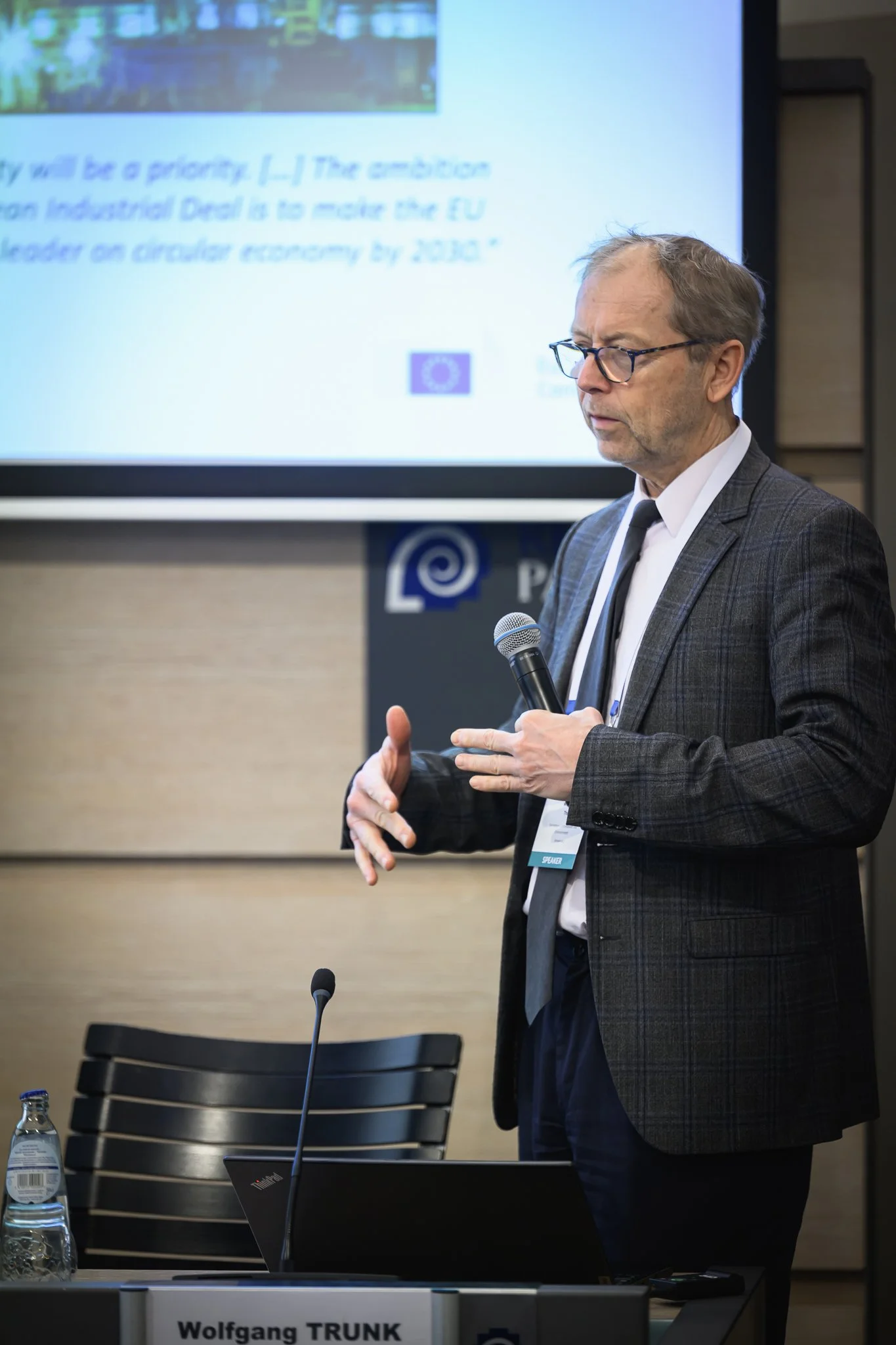 A man with glasses presents beside a screen in a smaller conference room.