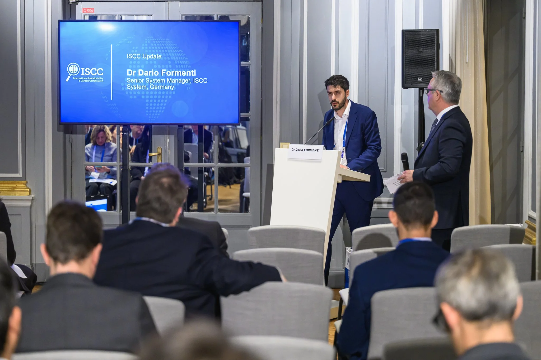 A speaker at a lectern addresses a smaller audience beside a digital presentation screen.