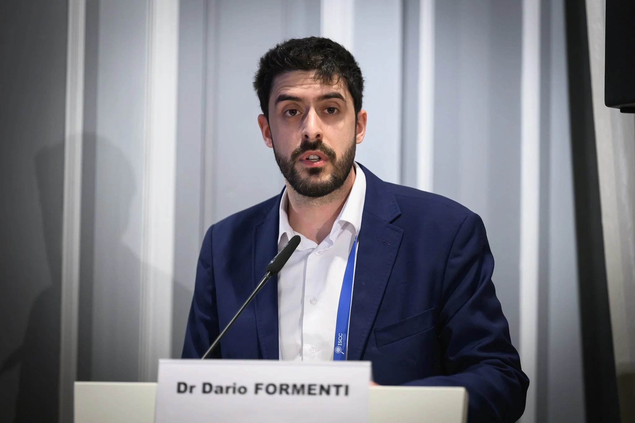 A man speaks at a lectern bearing the name Dr Dario Formenti.