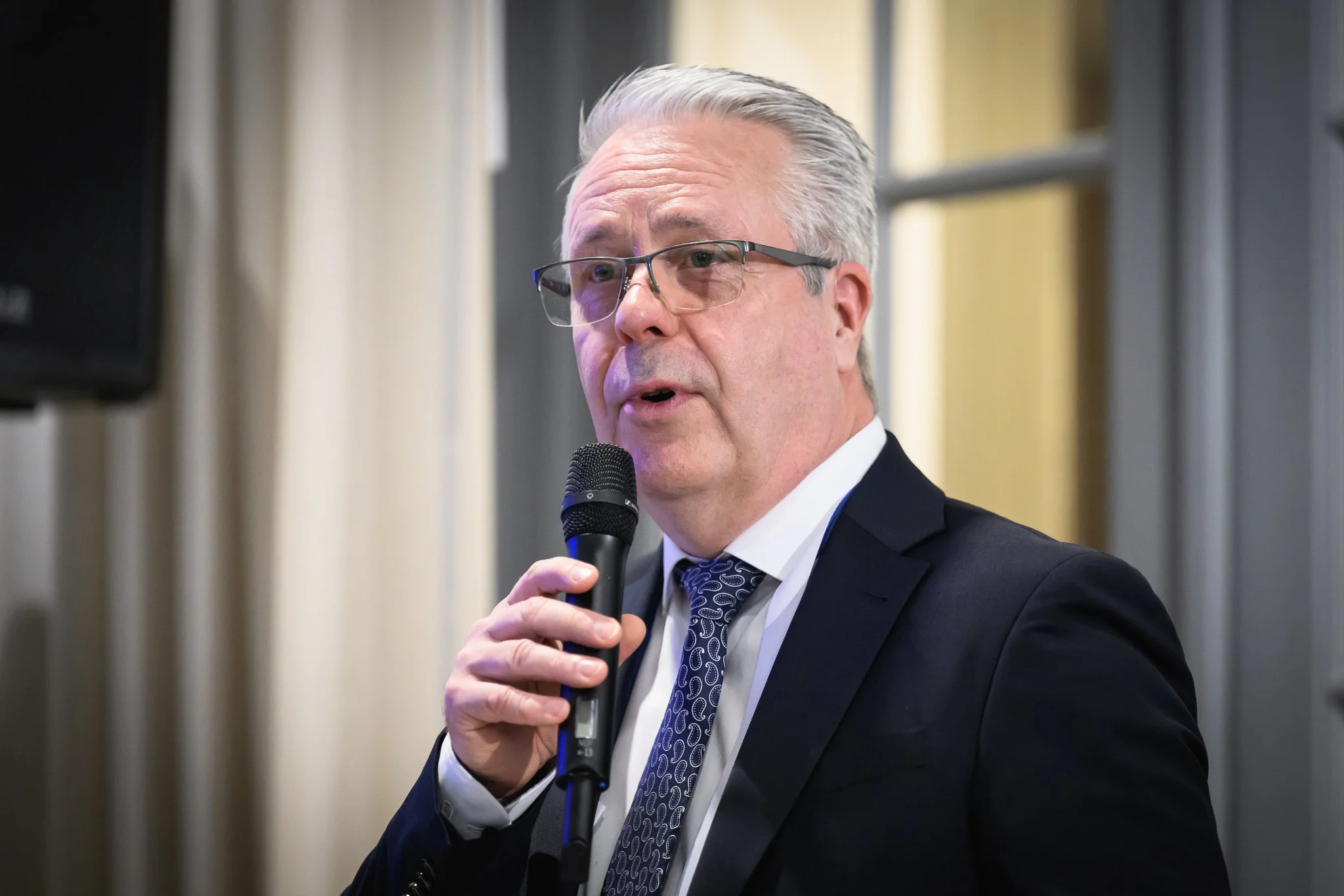 An older man with glasses speaks into a handheld microphone.