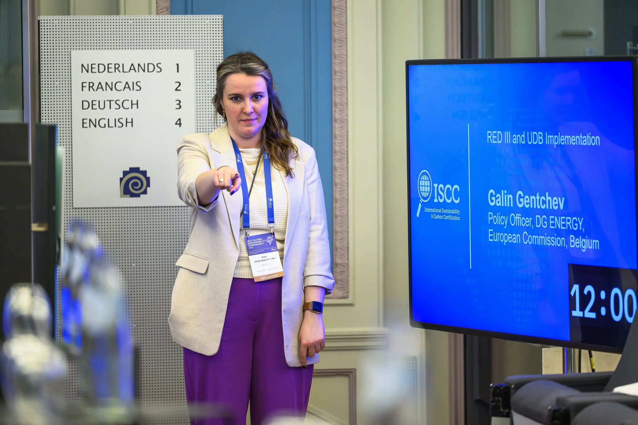 A woman wearing a headset gestures towards the audience beside a session information screen.