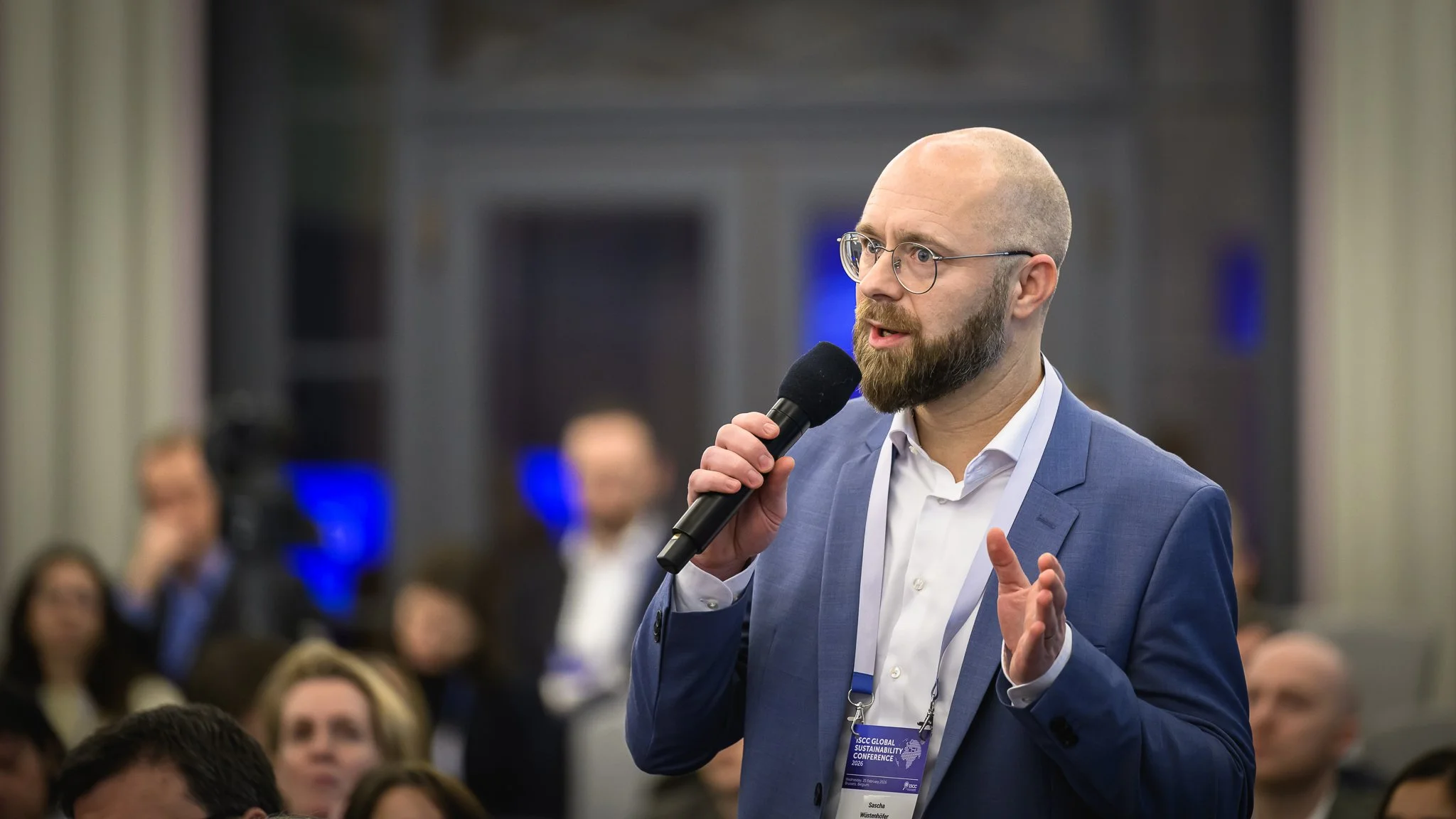 A bald man with glasses asks a question using a handheld microphone.