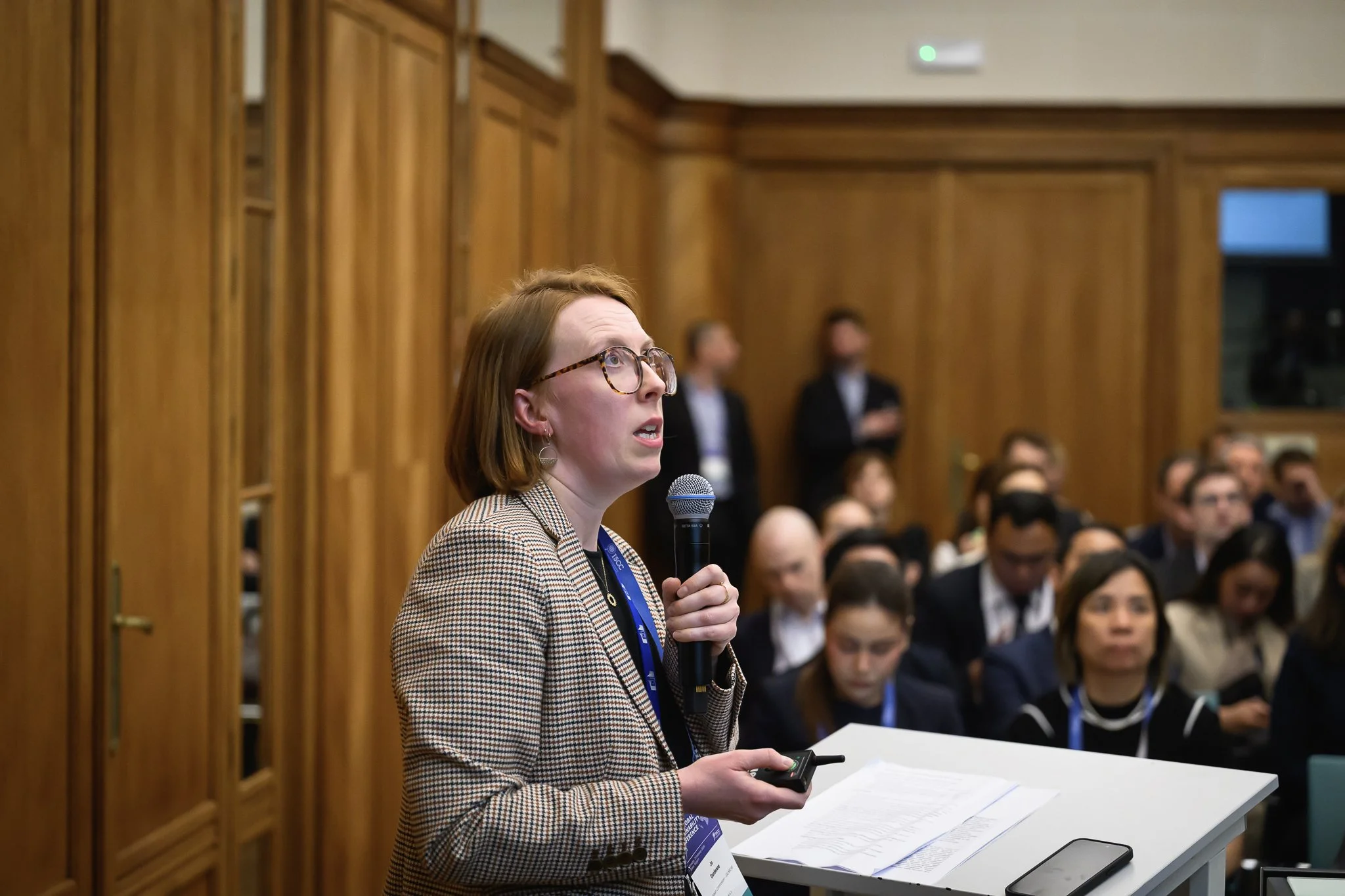 A woman with glasses speaks into a microphone beside a white lectern in a wood-panelled room.