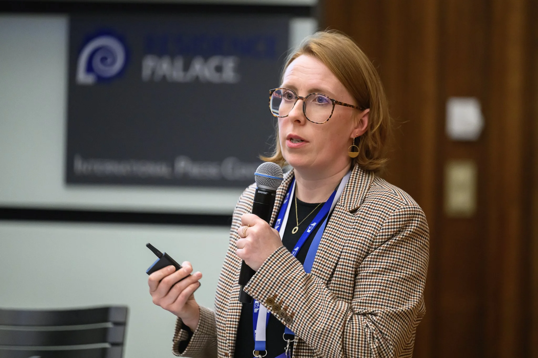 A woman with glasses speaks into a handheld microphone while holding a remote control.