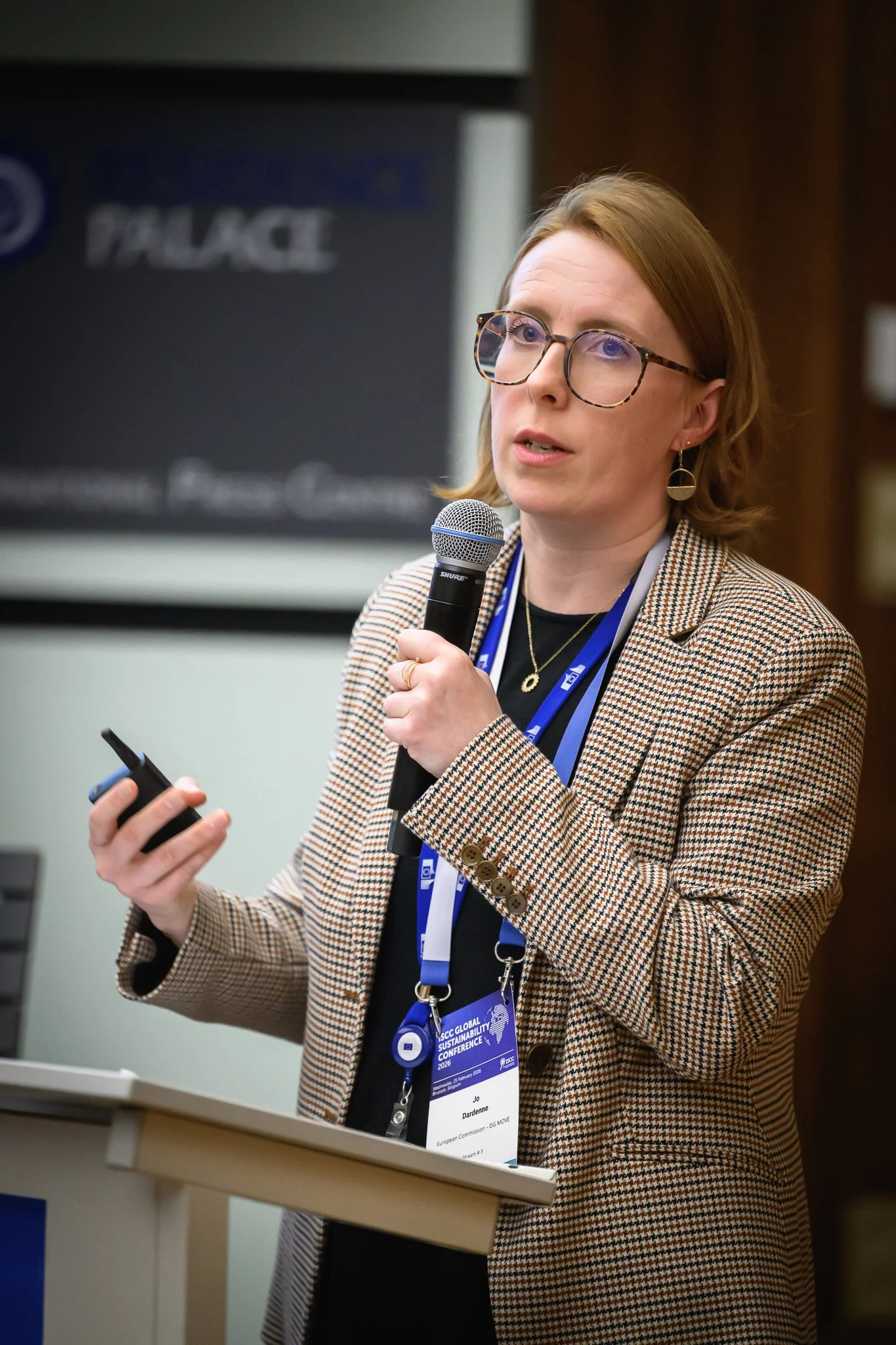 A woman with glasses presents while holding a microphone and a smartphone.