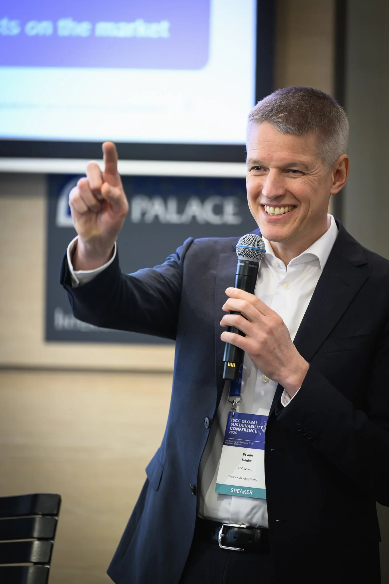 A smiling man holds a microphone and raises one finger while addressing the audience.