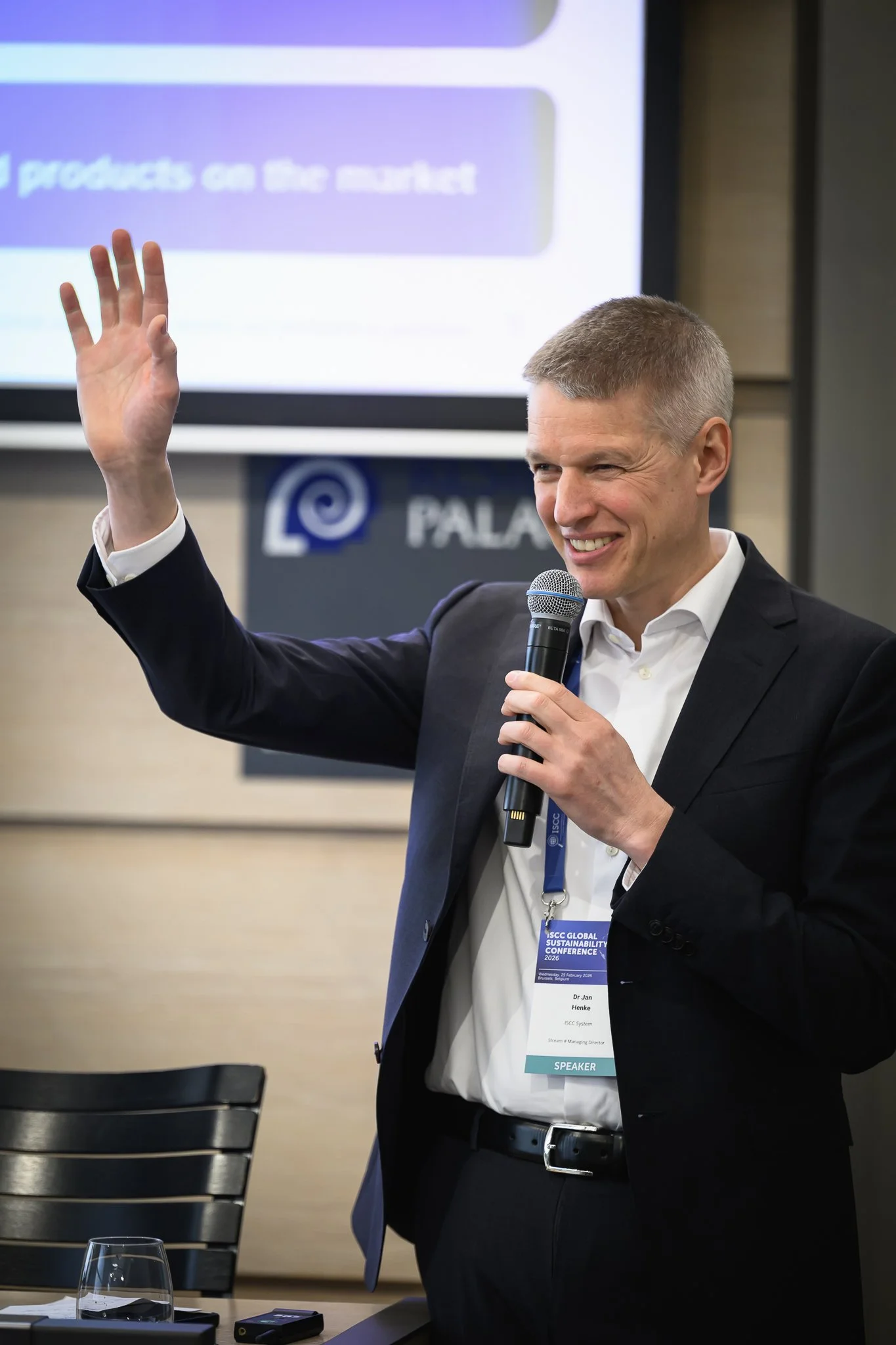 A smiling speaker raises one hand while holding a microphone in the other.