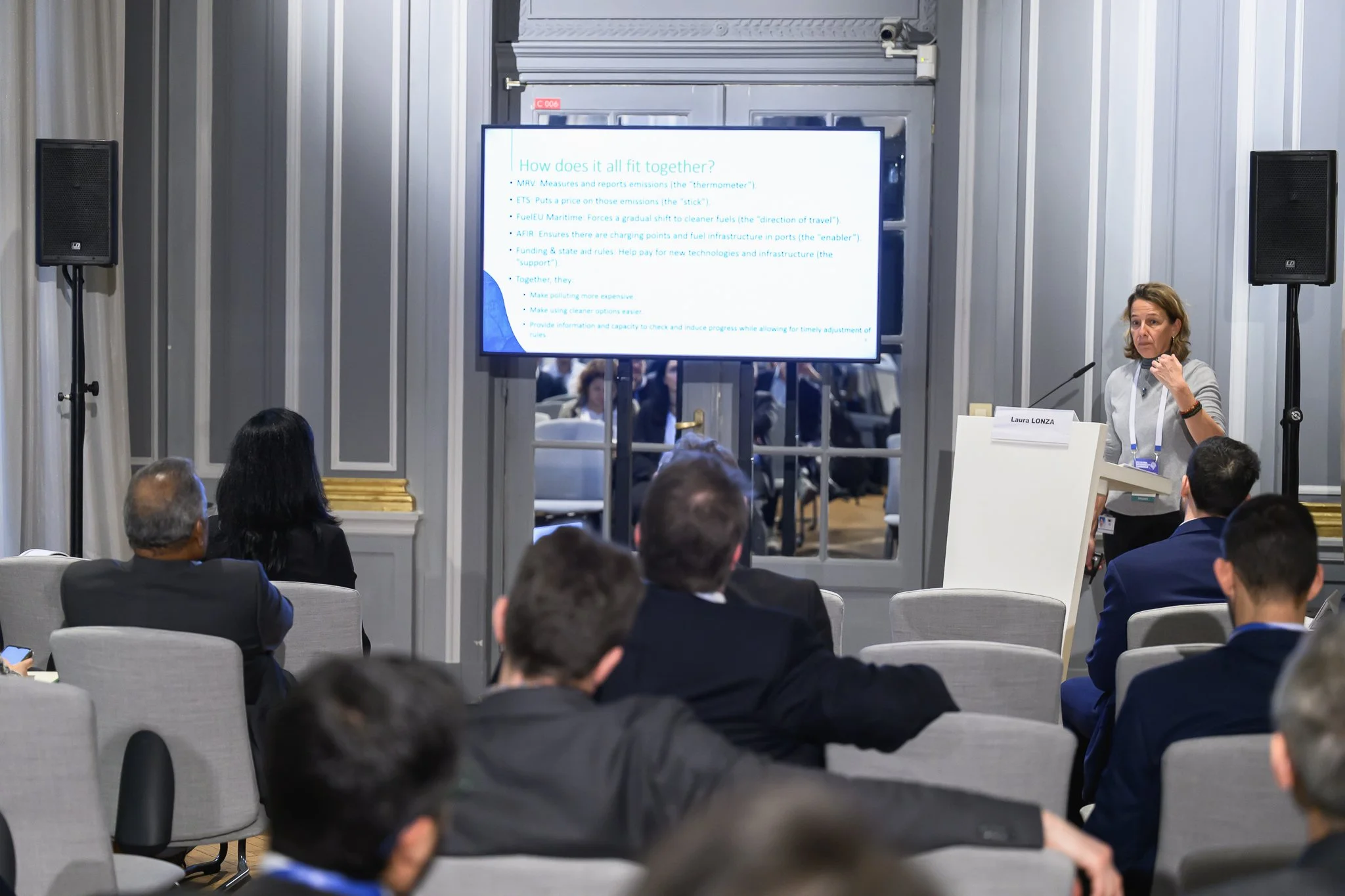 A seated audience watches a woman presenting beside a screen in a smaller conference room.