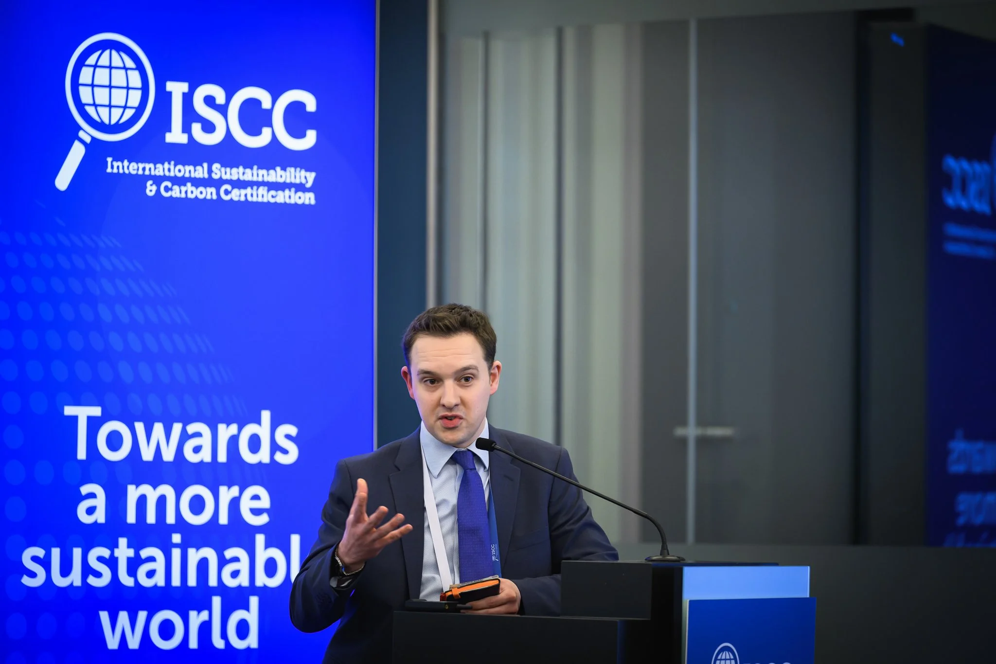 A man in a dark suit gestures at the lectern beside an ISCC banner.