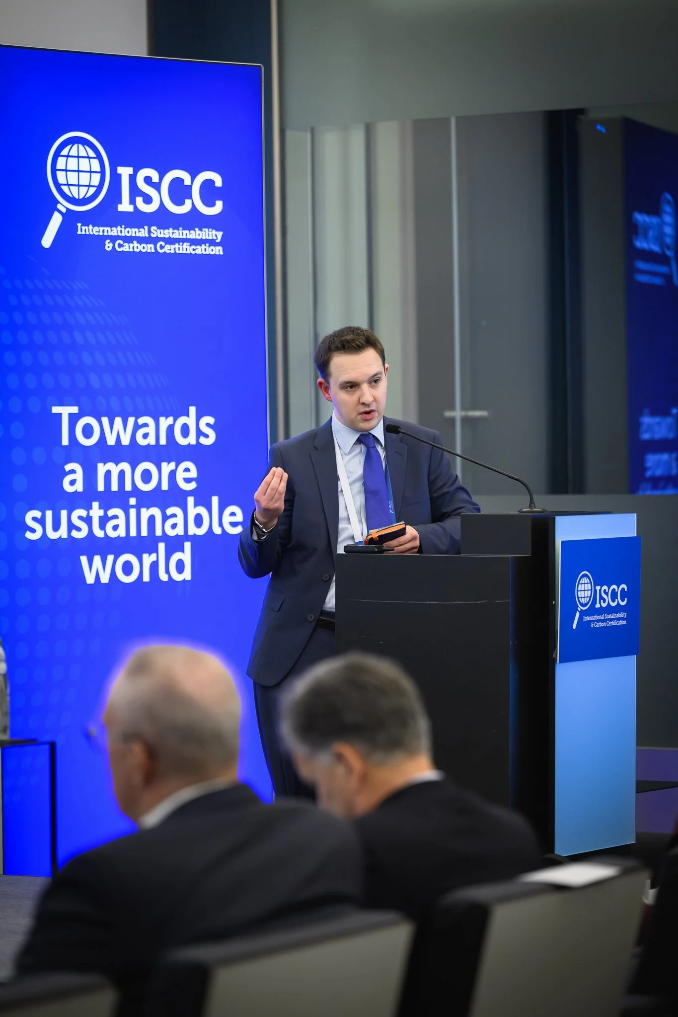A man presents from a lectern while conference participants listen.