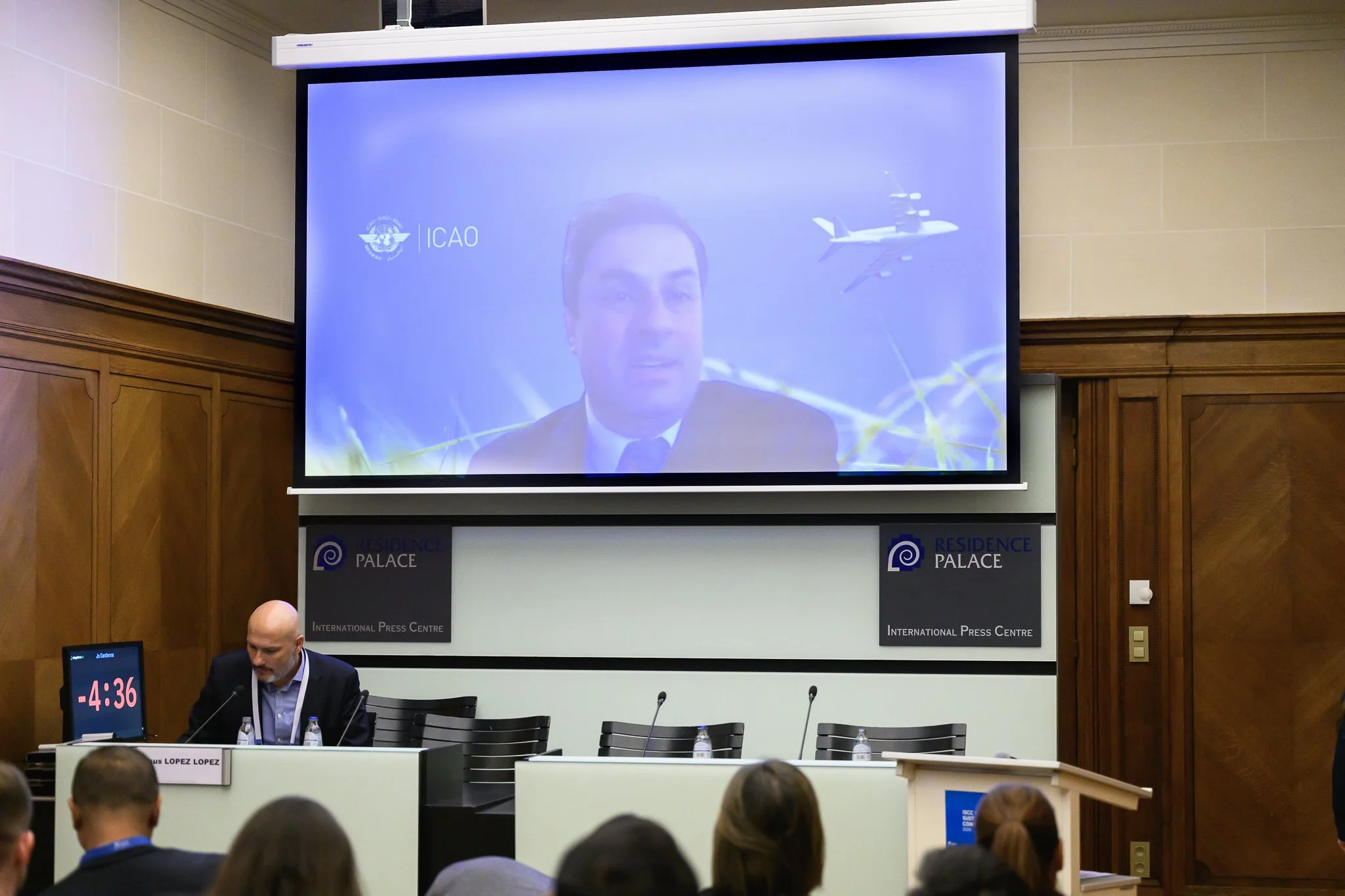 A remote speaker appears on a large screen above a seated participant in the conference room.