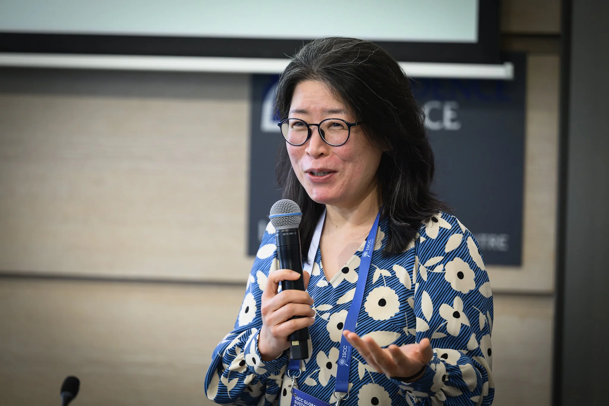 A woman with glasses and a patterned blouse speaks into a handheld microphone.
