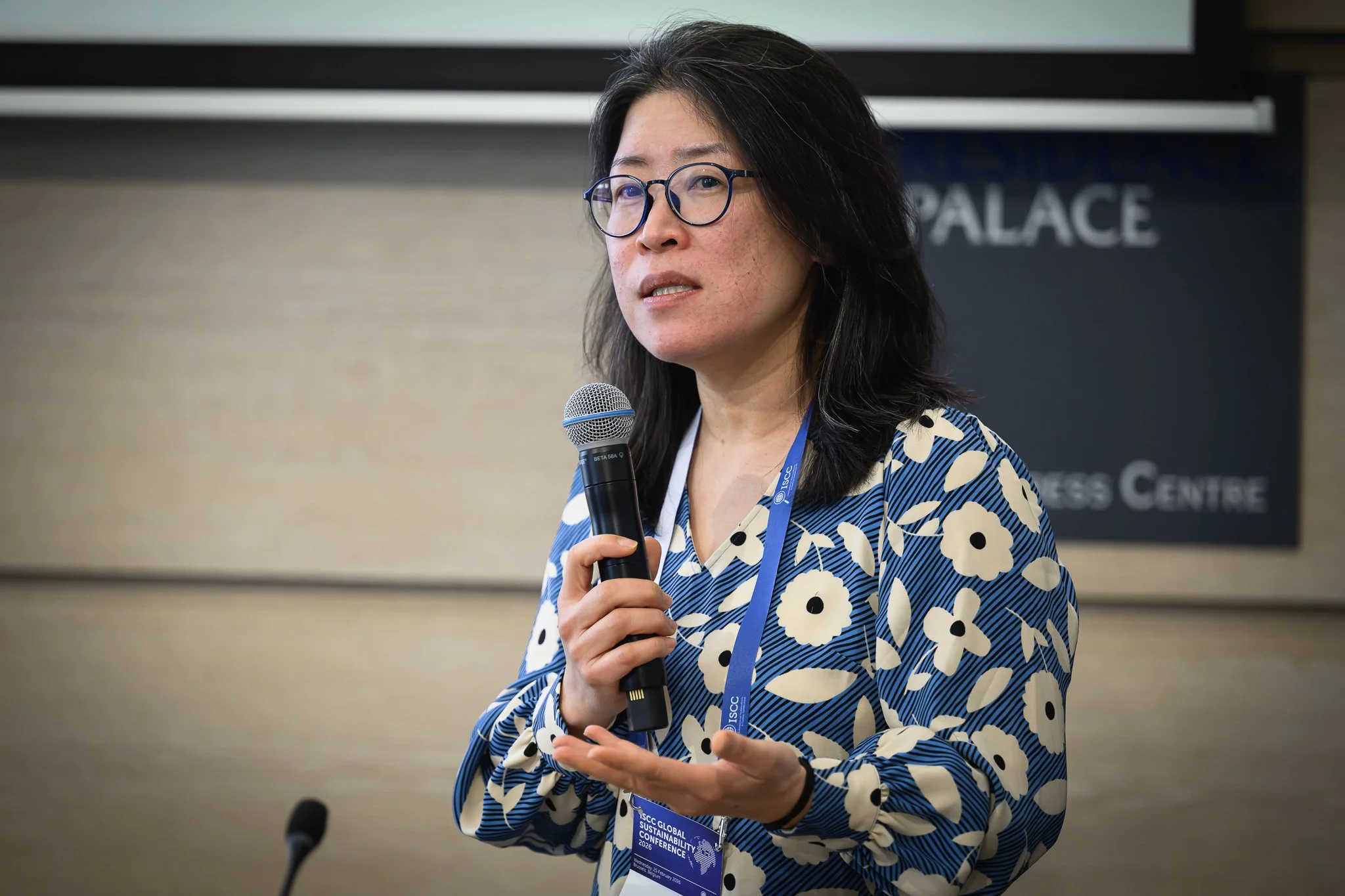 A woman with glasses speaks into a microphone and gestures with an open hand.
