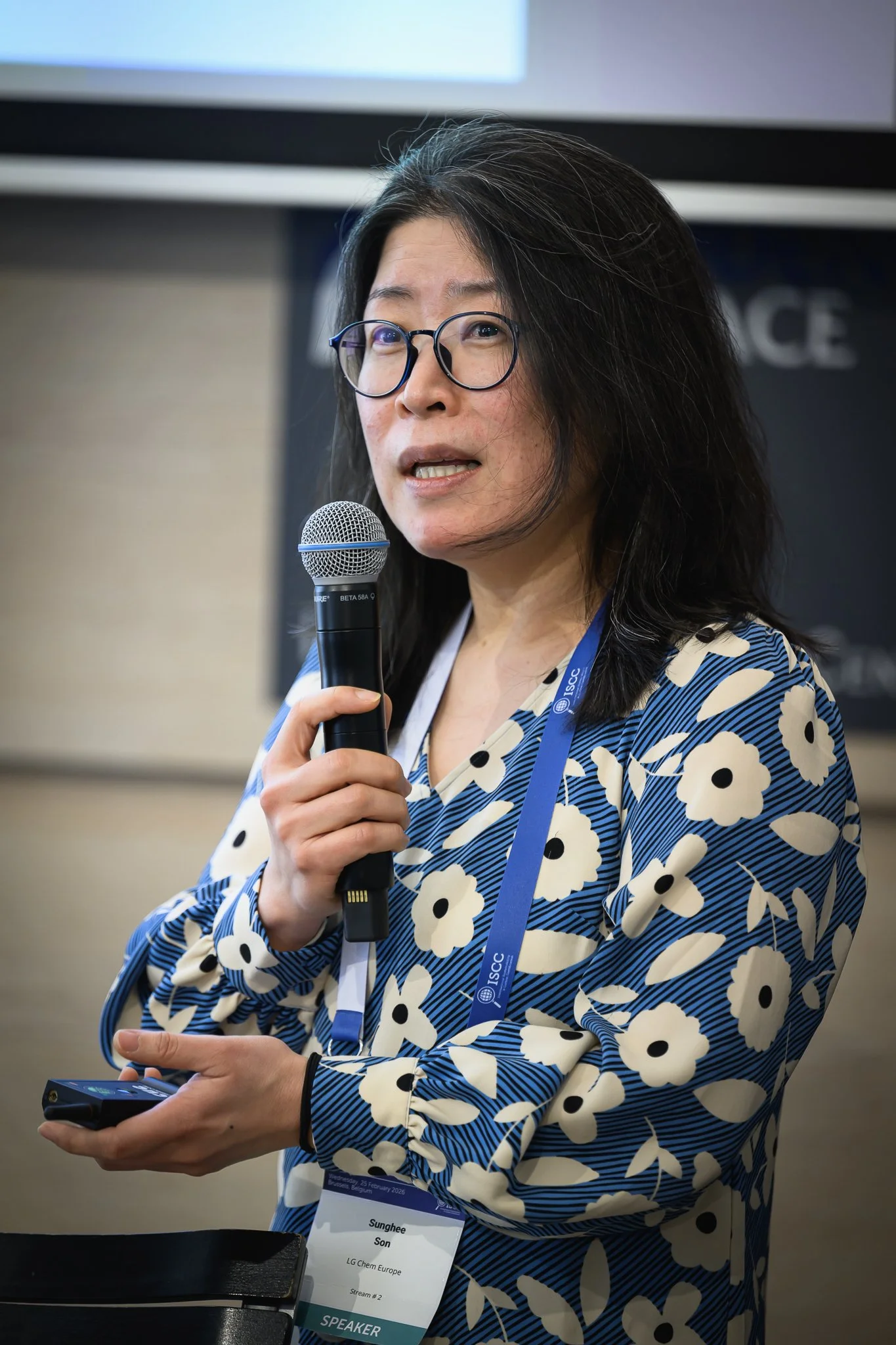 A woman in a patterned blouse gestures while holding a microphone and presentation remote.